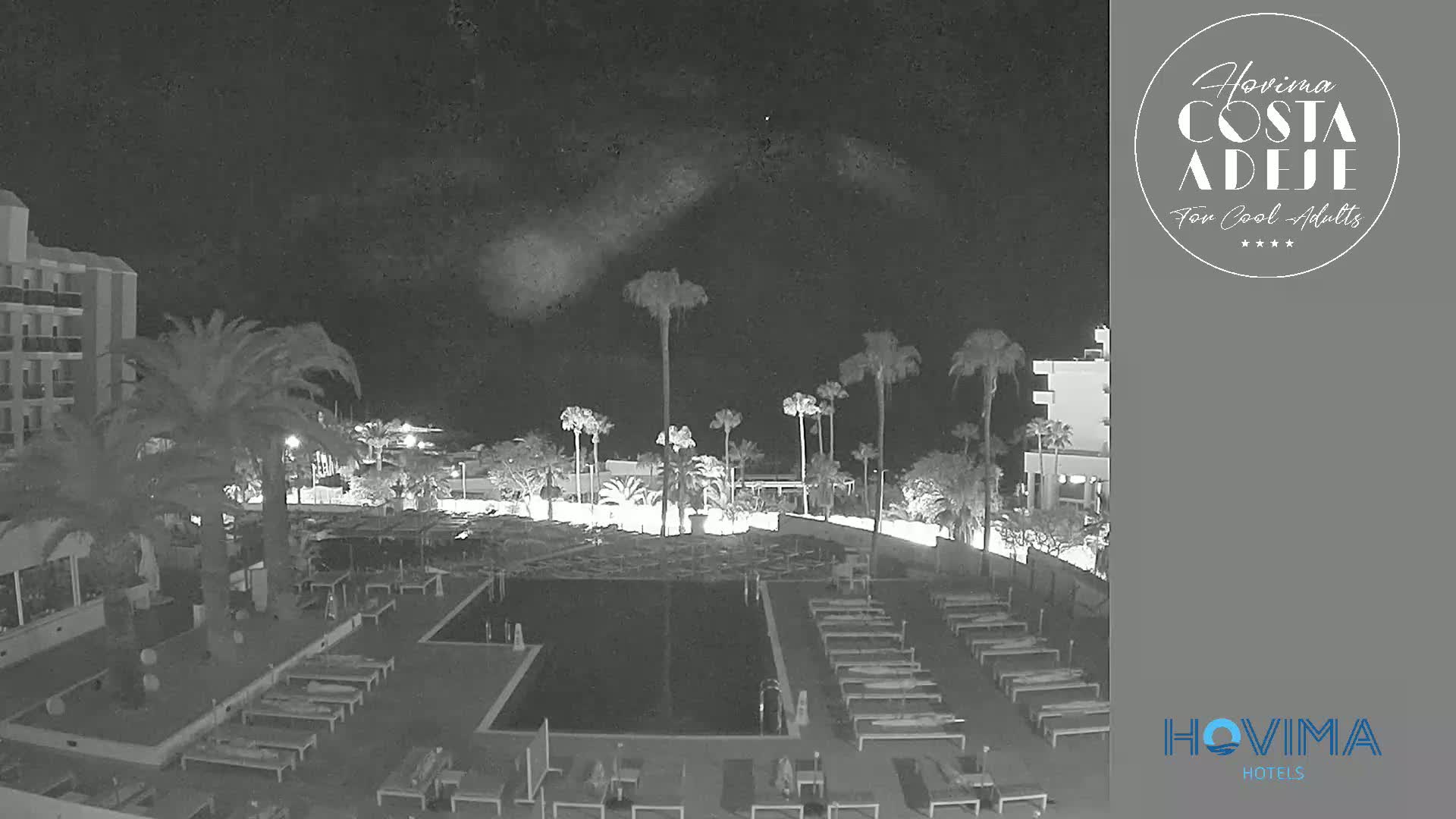 A nighttime view of a resort pool area with palm trees and a hotel building under a cloudy sky.