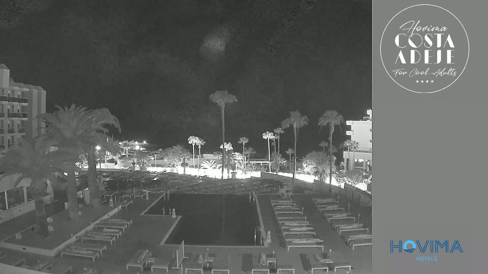 A nighttime, low-light view of a resort pool area with palm trees, lounge chairs, and a building in the background.
