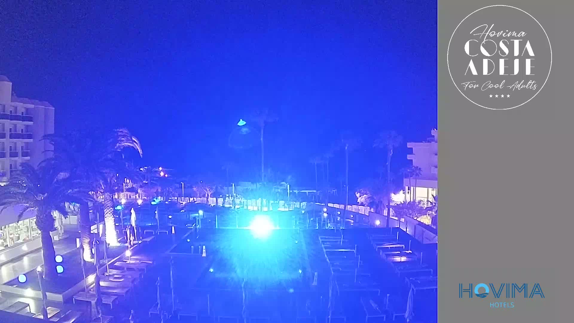 Under a dark blue night sky, a brightly lit resort pool area with lounge chairs and palm trees is visible near a multi-story hotel building.