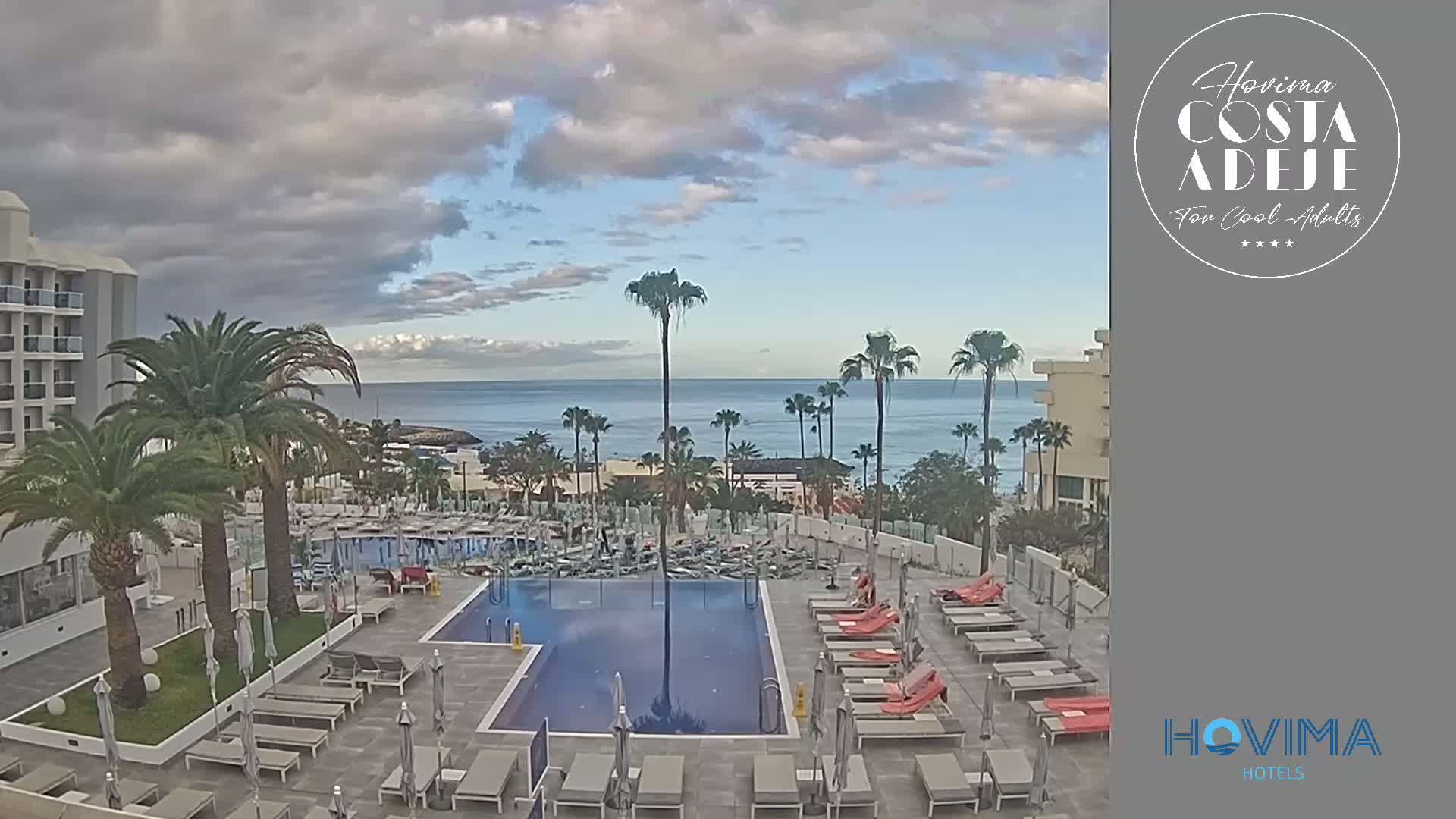 A resort pool area with lounge chairs and palm trees overlooks a calm ocean under a partly cloudy sky.