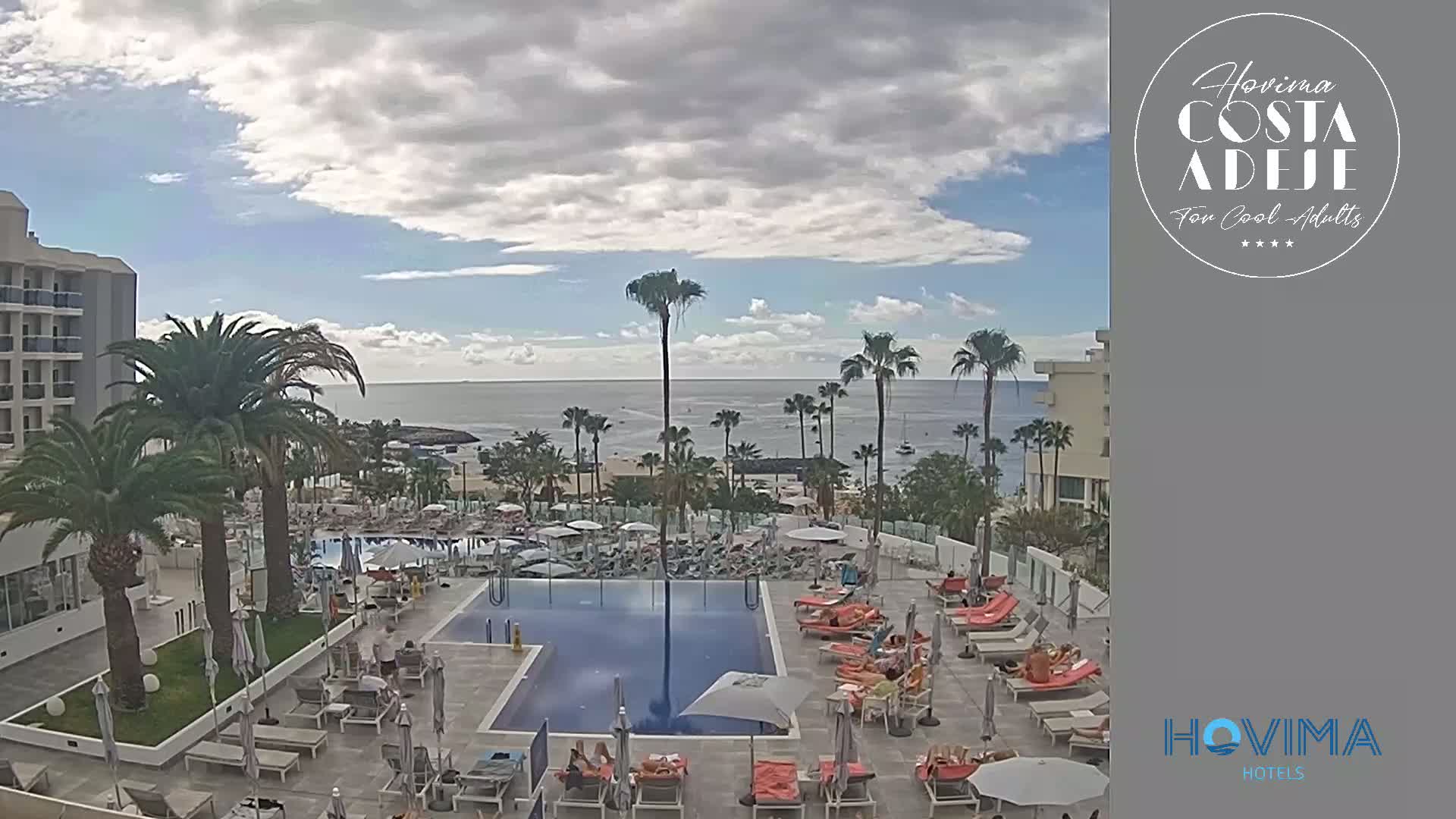 A partly cloudy day overlooks a resort pool area with palm trees and a beach beyond.