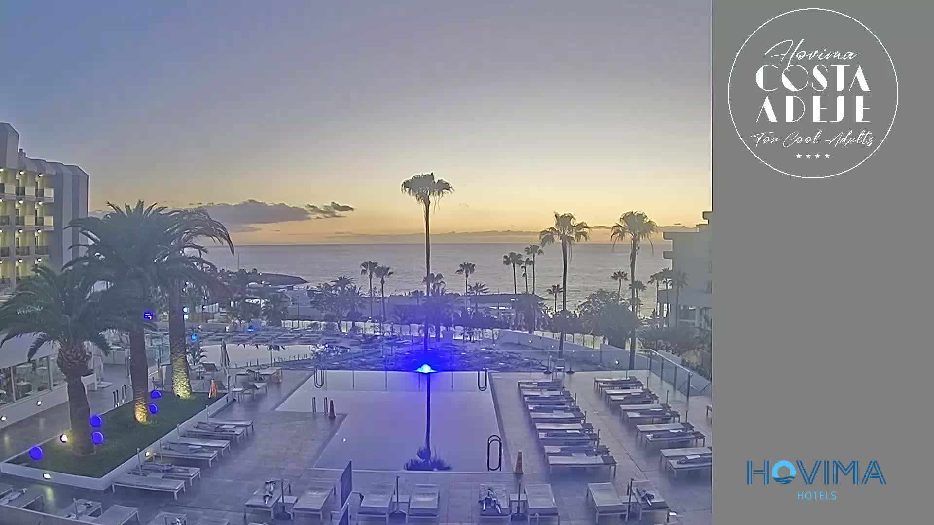 An outdoor pool area with lounge chairs sits beside a modern hotel building overlooking a calm ocean at sunset.