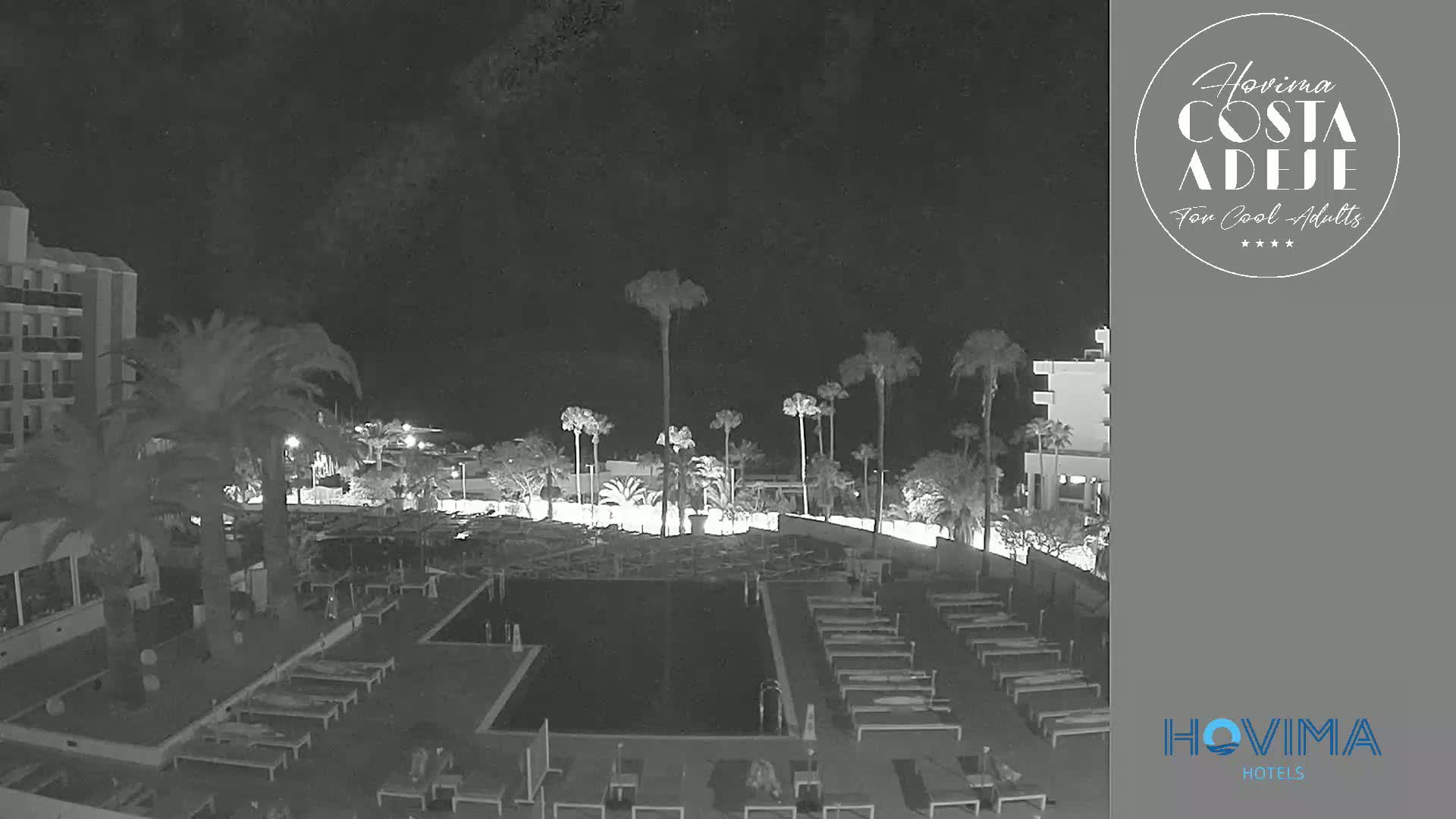 A nighttime view of a resort pool area with palm trees and buildings under a dark sky.