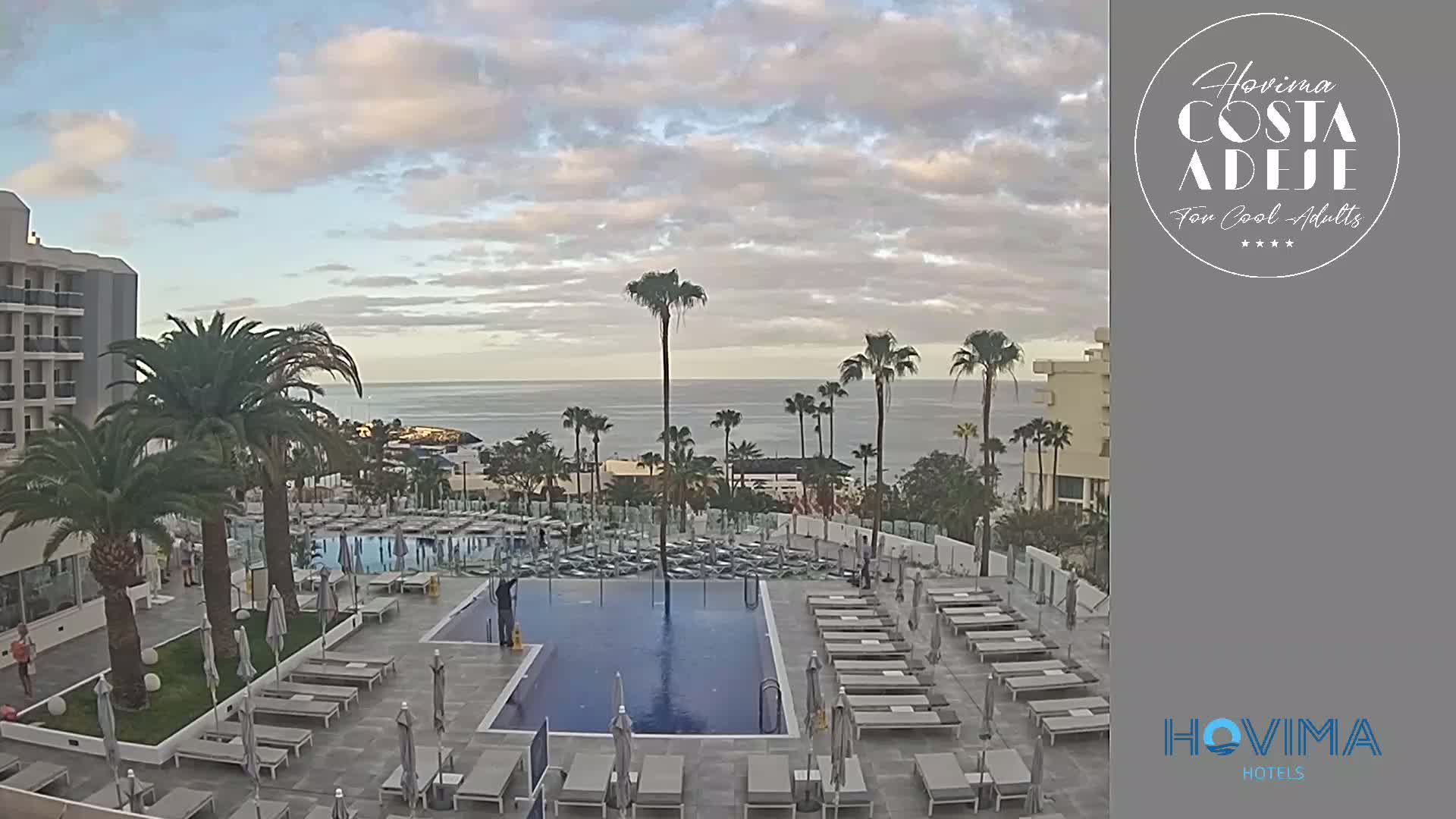 An outdoor pool area with numerous lounge chairs surrounding a rectangular pool, situated next to a hotel with palm trees and a calm ocean in the background under a cloudy sky.