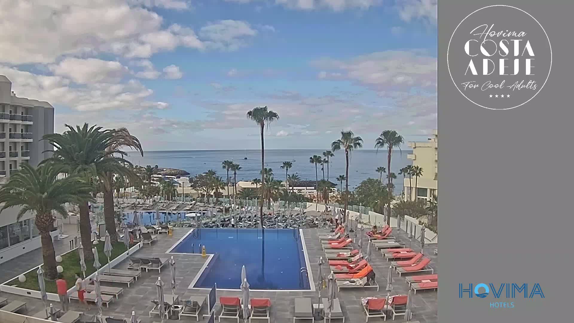 A resort pool area with orange lounge chairs and palm trees overlooks a calm ocean under a partly cloudy sky.