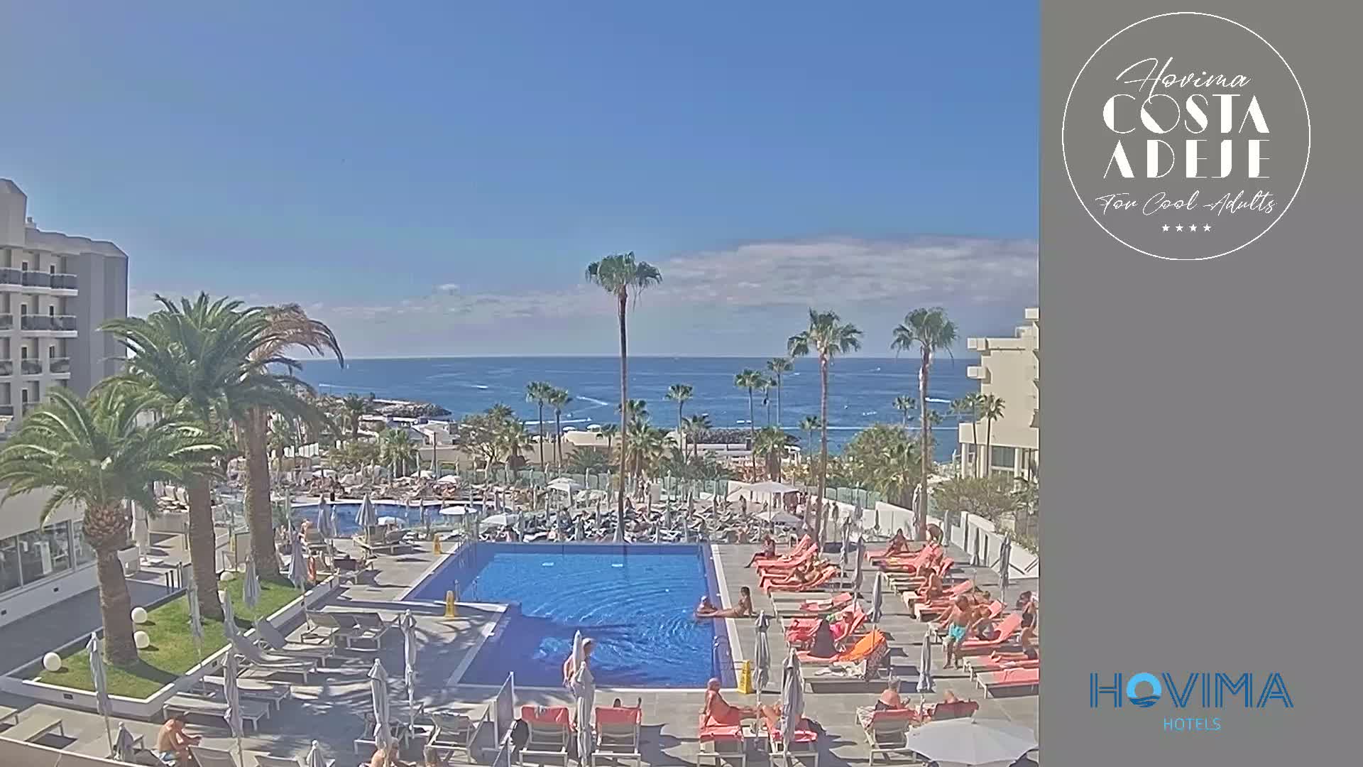 A resort pool area with many people relaxing on lounge chairs surrounds a swimming pool, all overlooking a calm ocean under a clear blue sky.