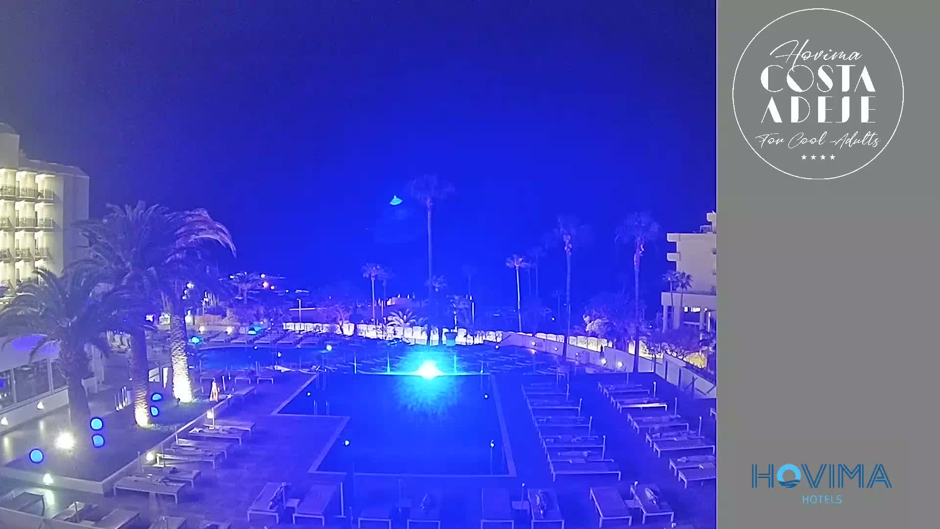 A nighttime view of a resort pool area, illuminated in blue, surrounded by palm trees and a hotel building under a clear, dark blue sky.
