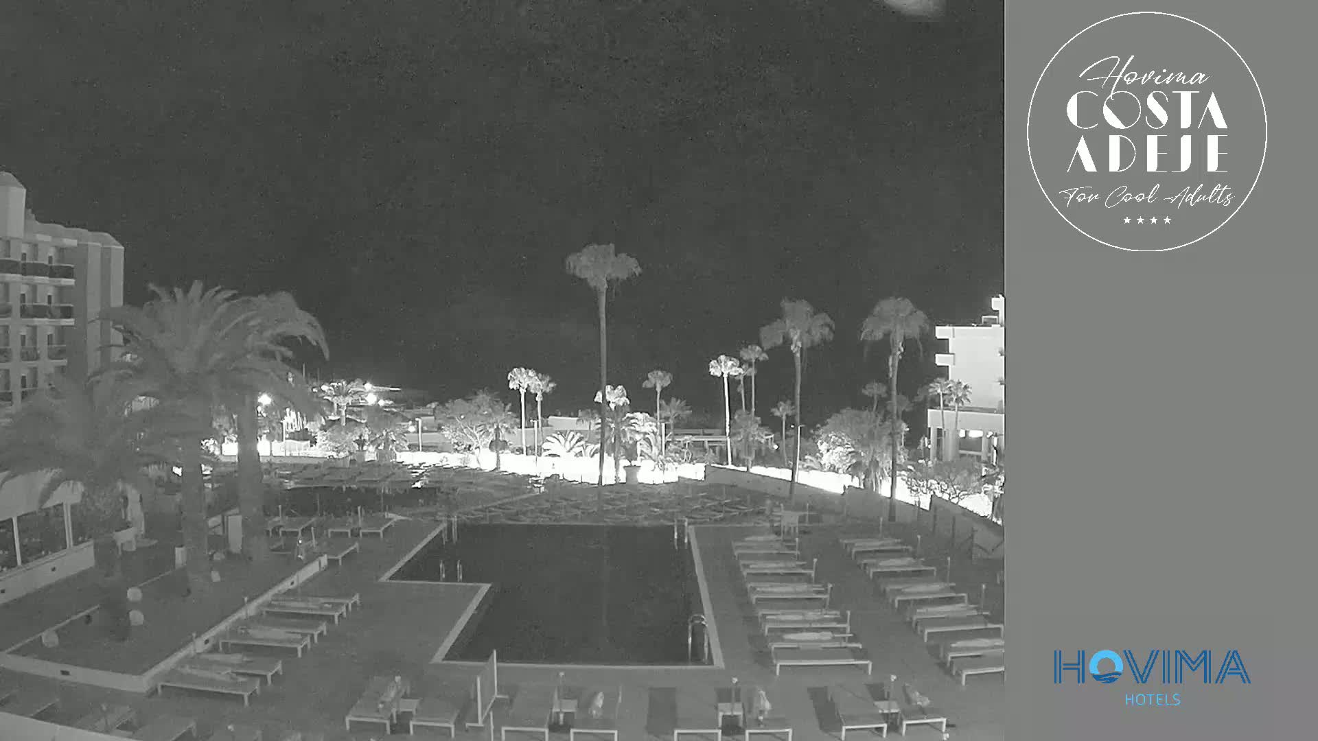 A nighttime view of a resort pool area with palm trees, lounge chairs, and a hotel building under a dark, clear sky.