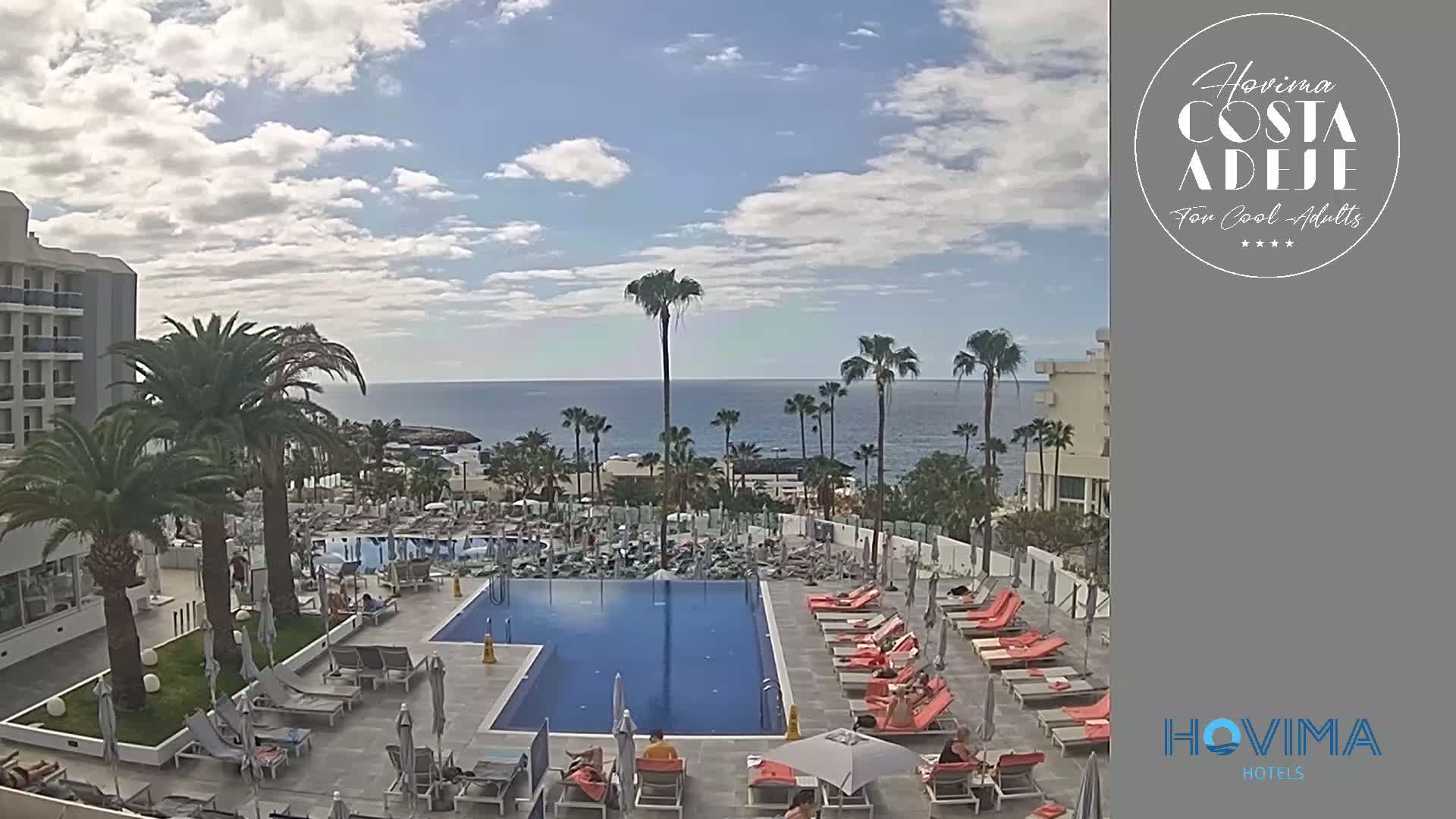A resort pool area with lounge chairs and palm trees overlooks a calm ocean under a partly cloudy sky.