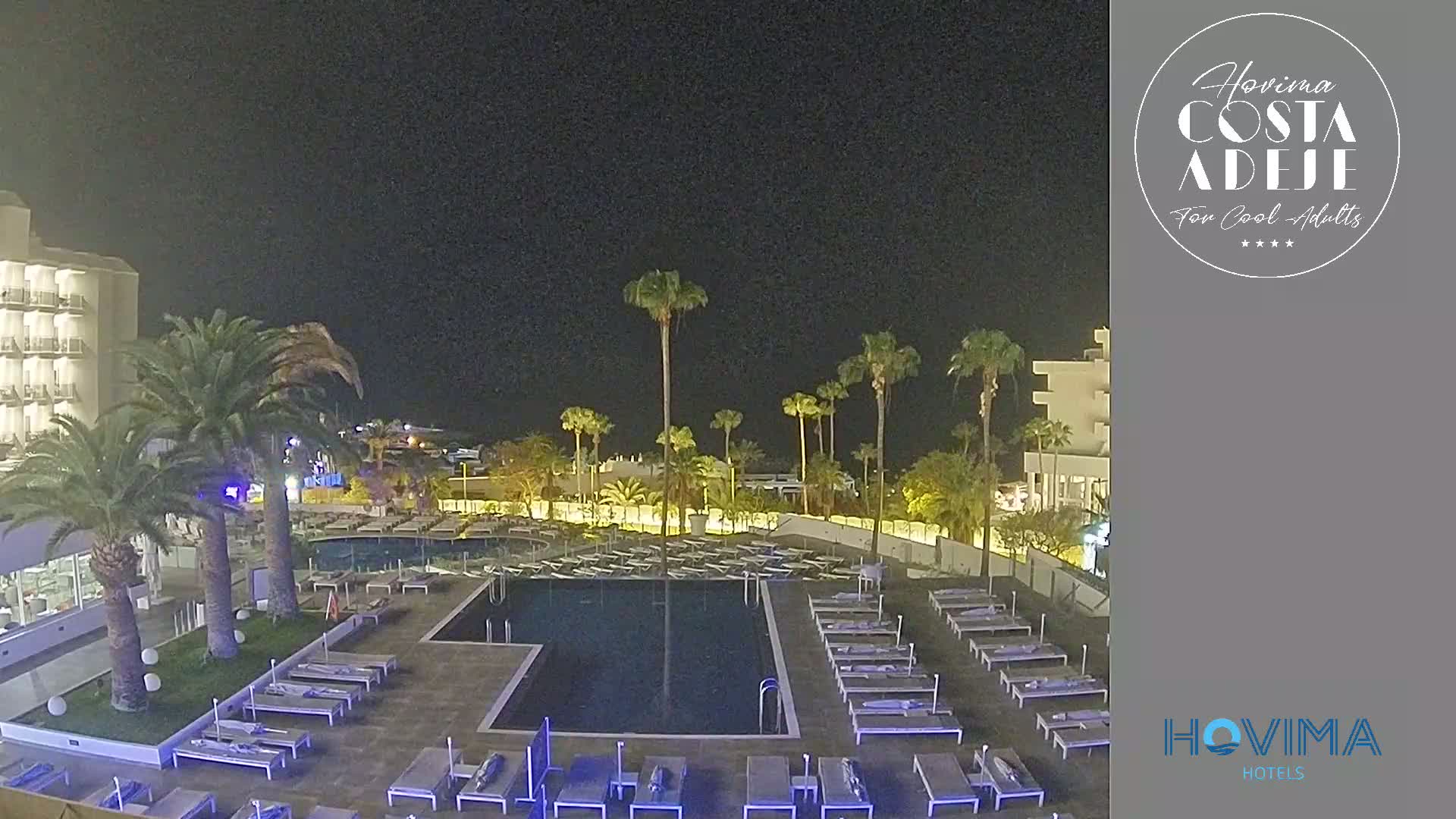 A nighttime view of a resort pool area with palm trees, lounge chairs, and a hotel building under a clear, dark sky.