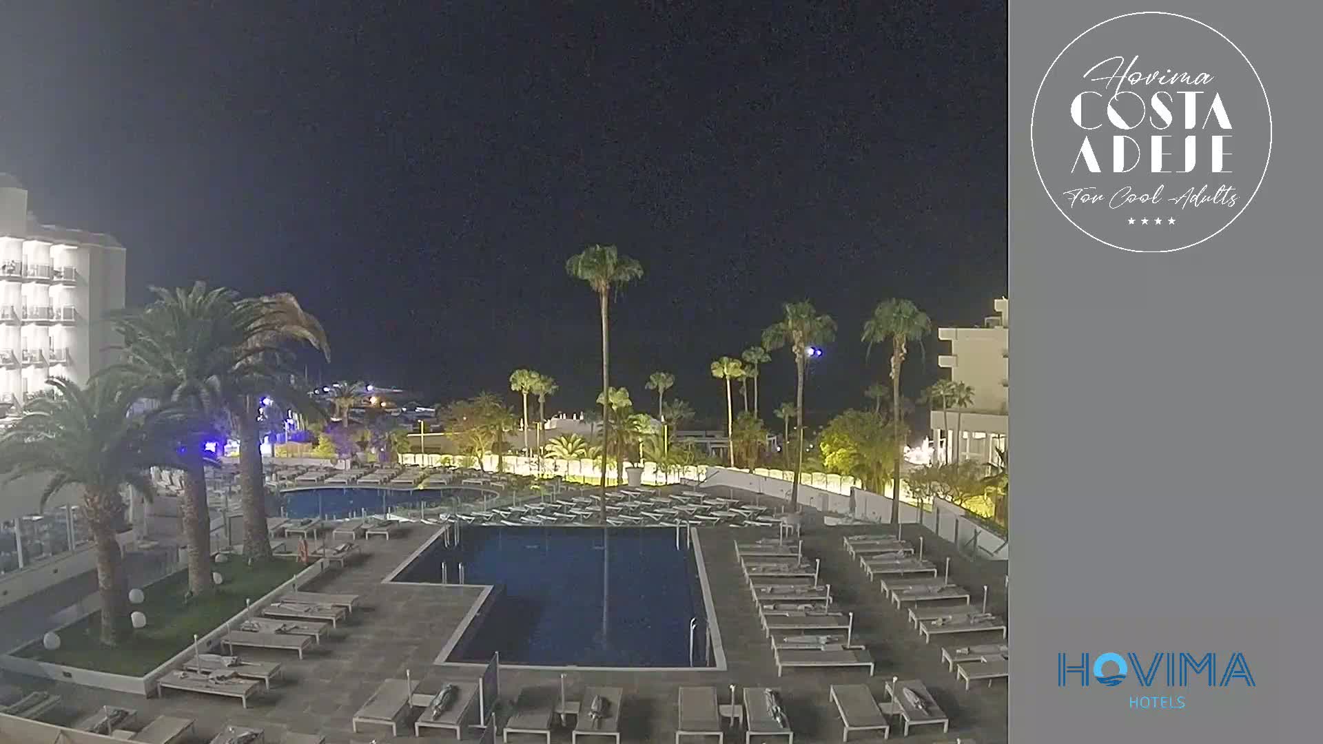 A nighttime view of a resort pool area with palm trees, lounge chairs, and a hotel building under a clear, dark sky.