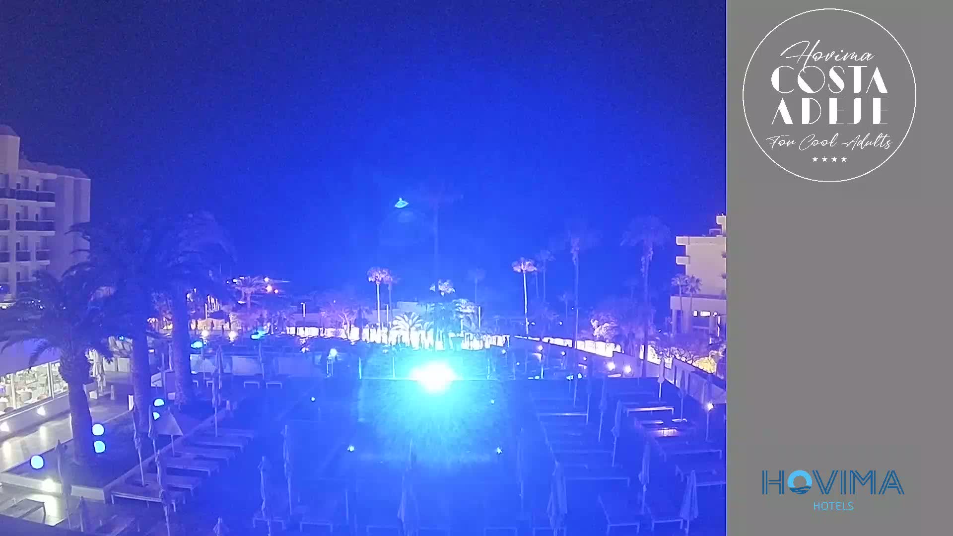 A nighttime view of a resort pool area with palm trees and buildings illuminated in blue light.