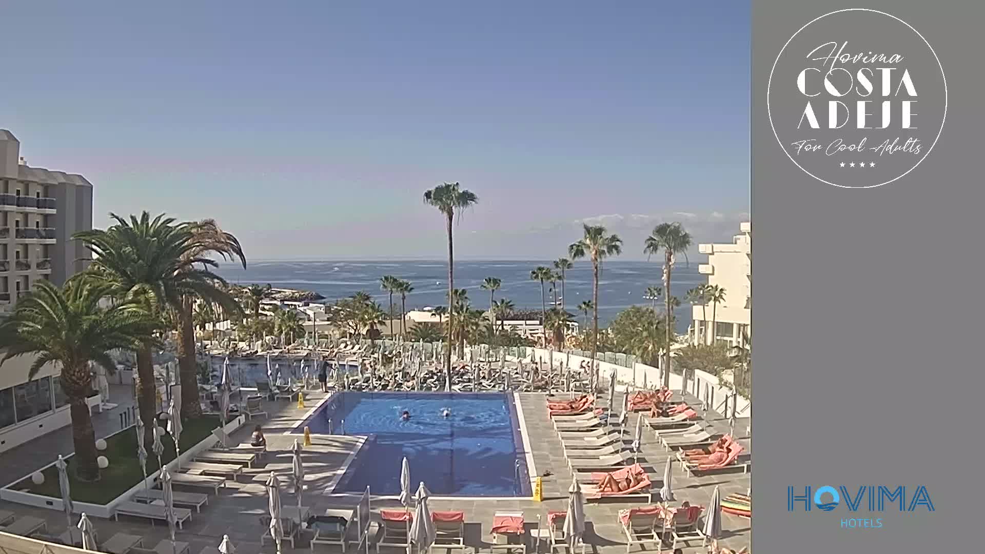 A resort pool area with sunbathers and swimmers is situated next to a hotel overlooking a calm ocean under a clear blue sky.