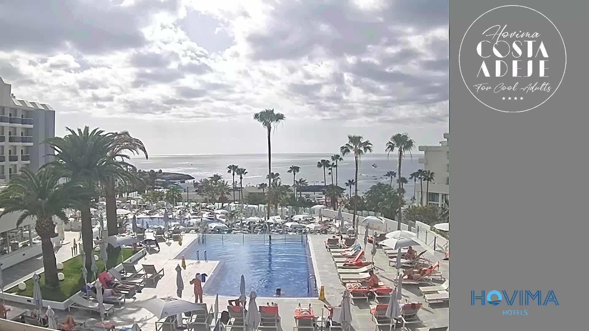 A resort pool area with lounge chairs and umbrellas overlooks a calm ocean under a partly cloudy sky.