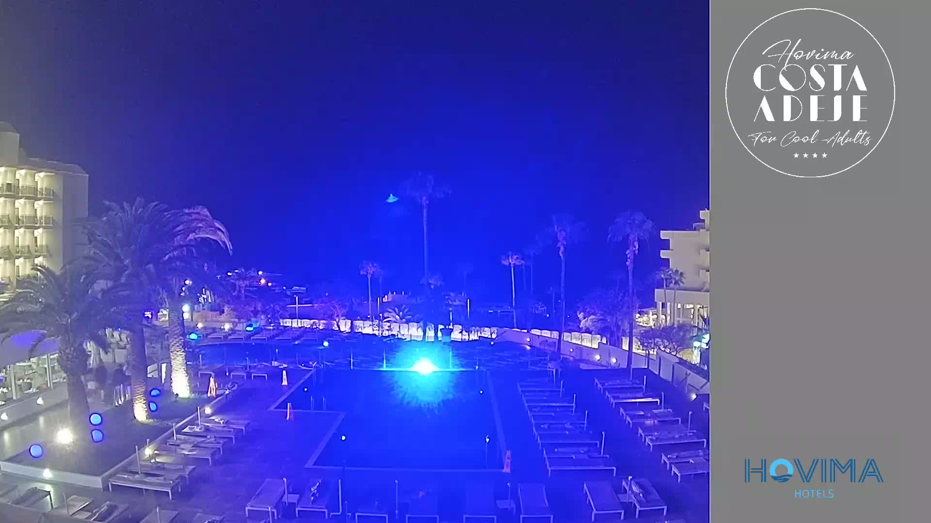A nighttime view of a resort pool area with palm trees and a hotel building under a dark sky.