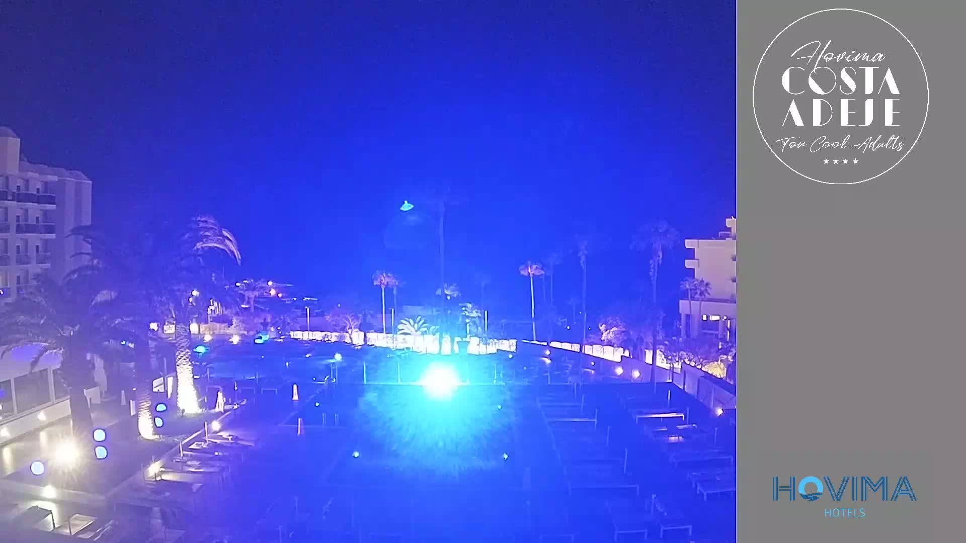 A nighttime view of a resort pool area, illuminated in blue light, with palm trees and buildings surrounding it under a clear, dark sky.