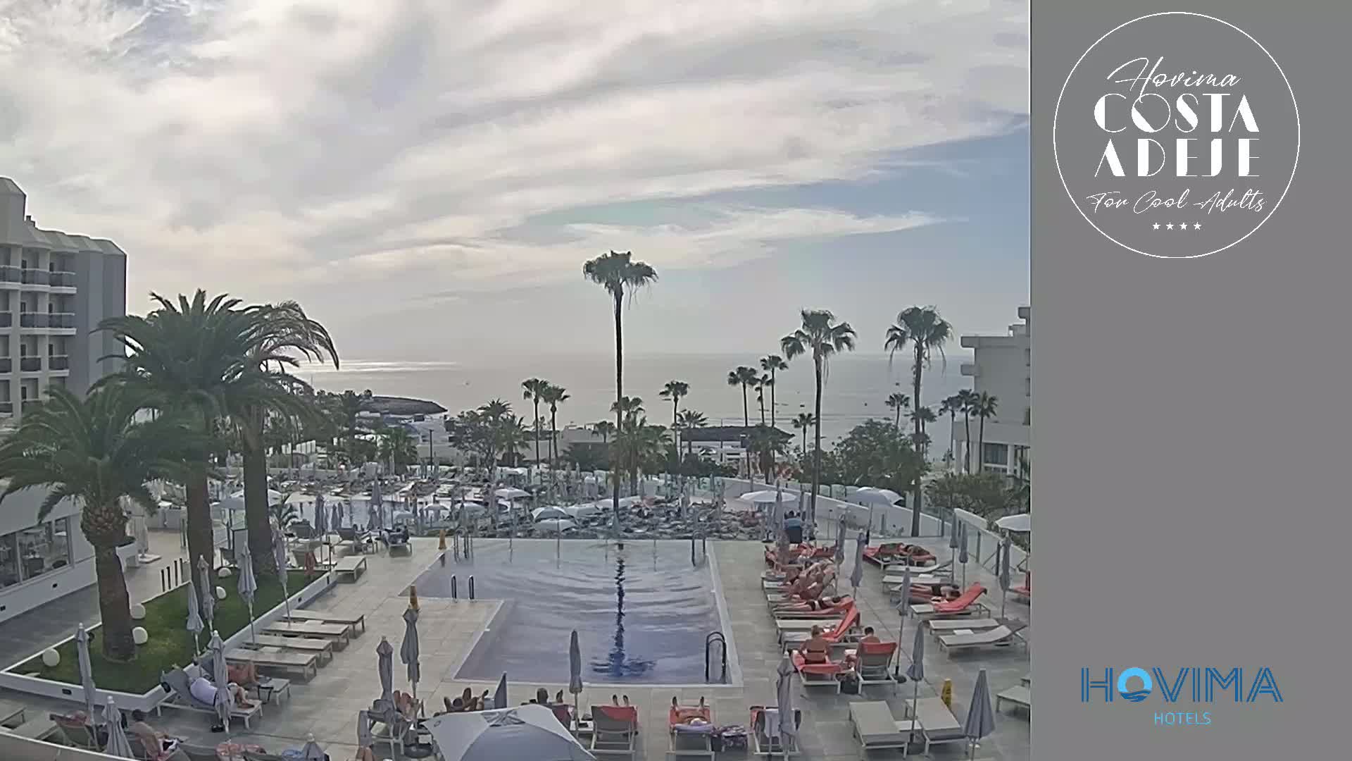 An outdoor pool area with lounge chairs and people relaxing, situated next to a large hotel with palm trees and an ocean view under a mostly cloudy sky.