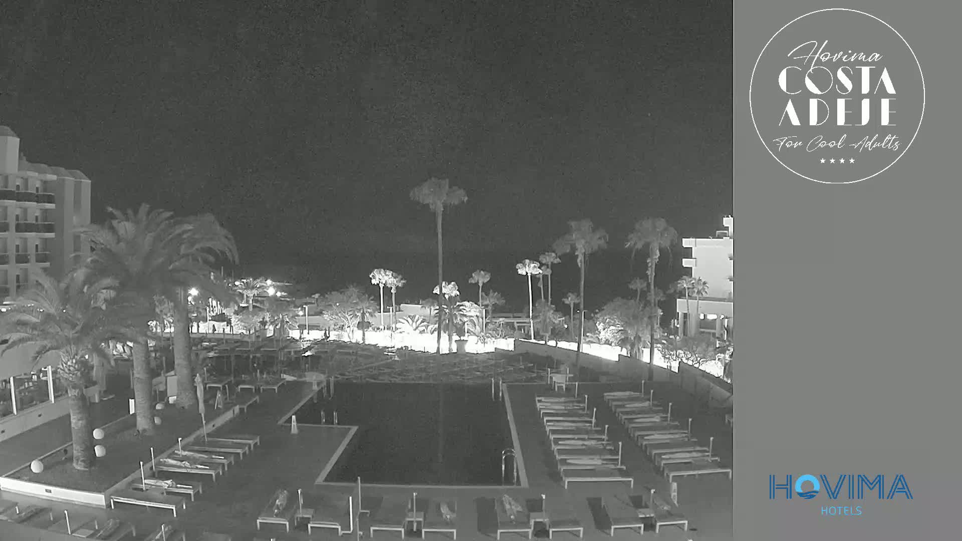 A nighttime, low-light view of a resort pool area with palm trees and buildings surrounding it.