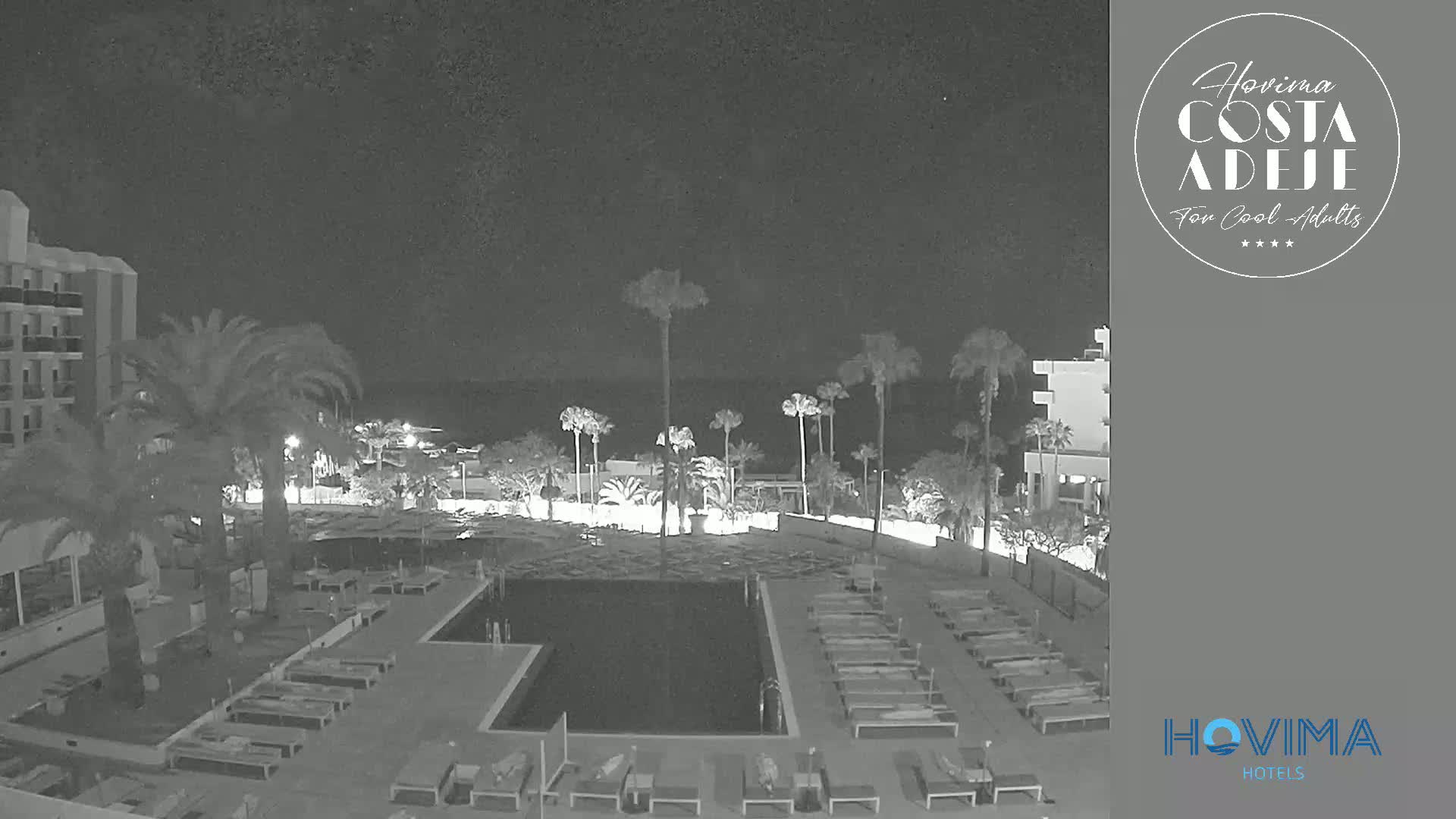 A nighttime view of a resort pool area with palm trees, lounge chairs, and a building under a clear, dark sky.