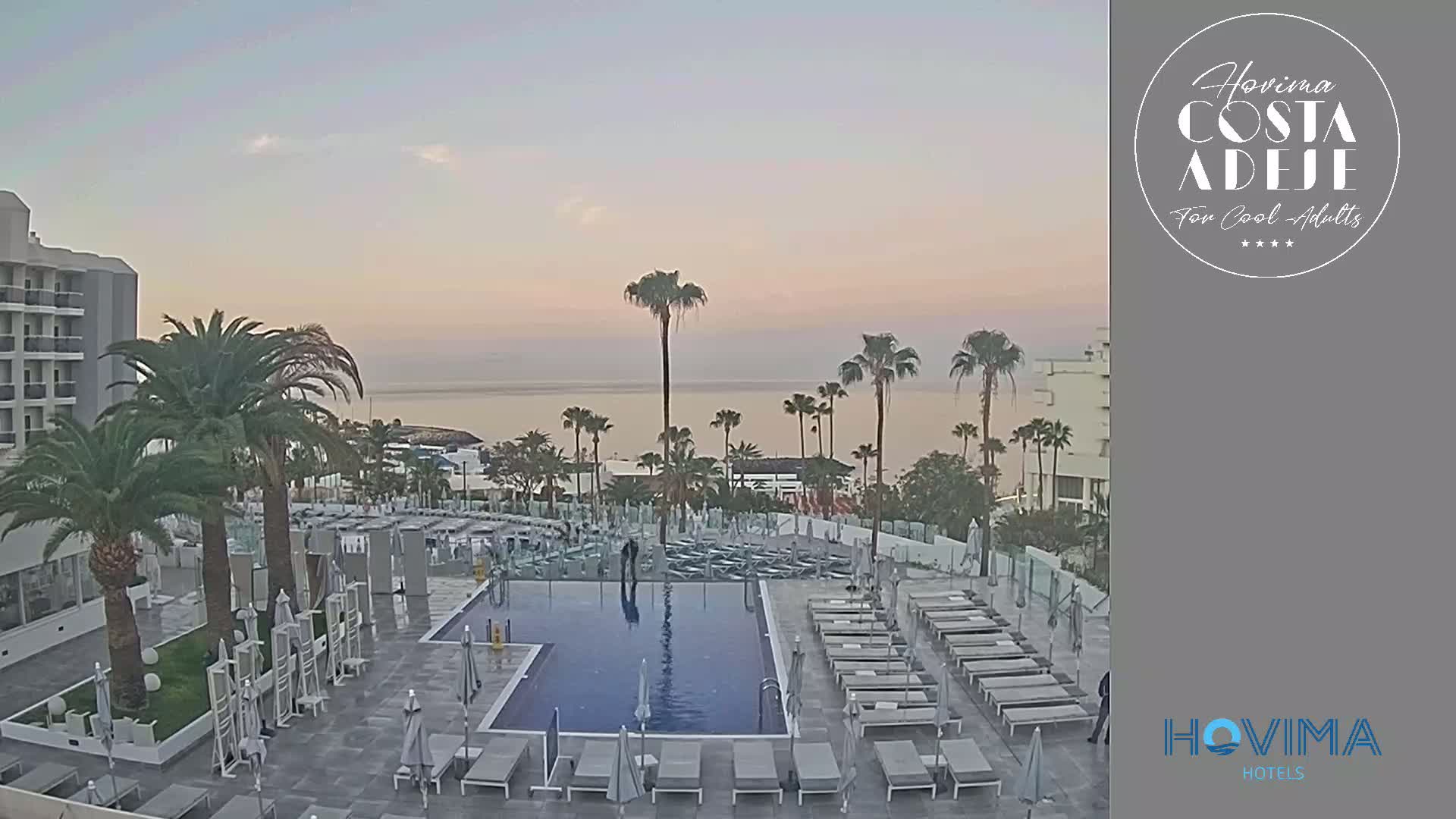 A resort pool area with lounge chairs surrounds a rectangular pool, overlooking a calm ocean under a pale sunrise sky.