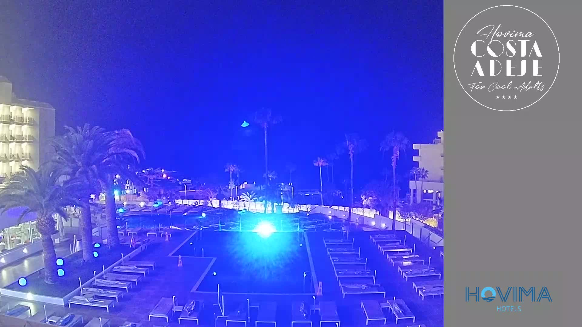 A nighttime view of a hotel pool area illuminated in blue light, surrounded by palm trees and buildings under a clear, dark blue sky.