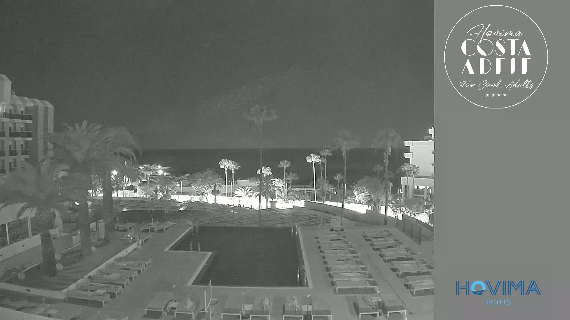 A nighttime view of a resort pool area with palm trees, buildings, and a calm ocean in the background.