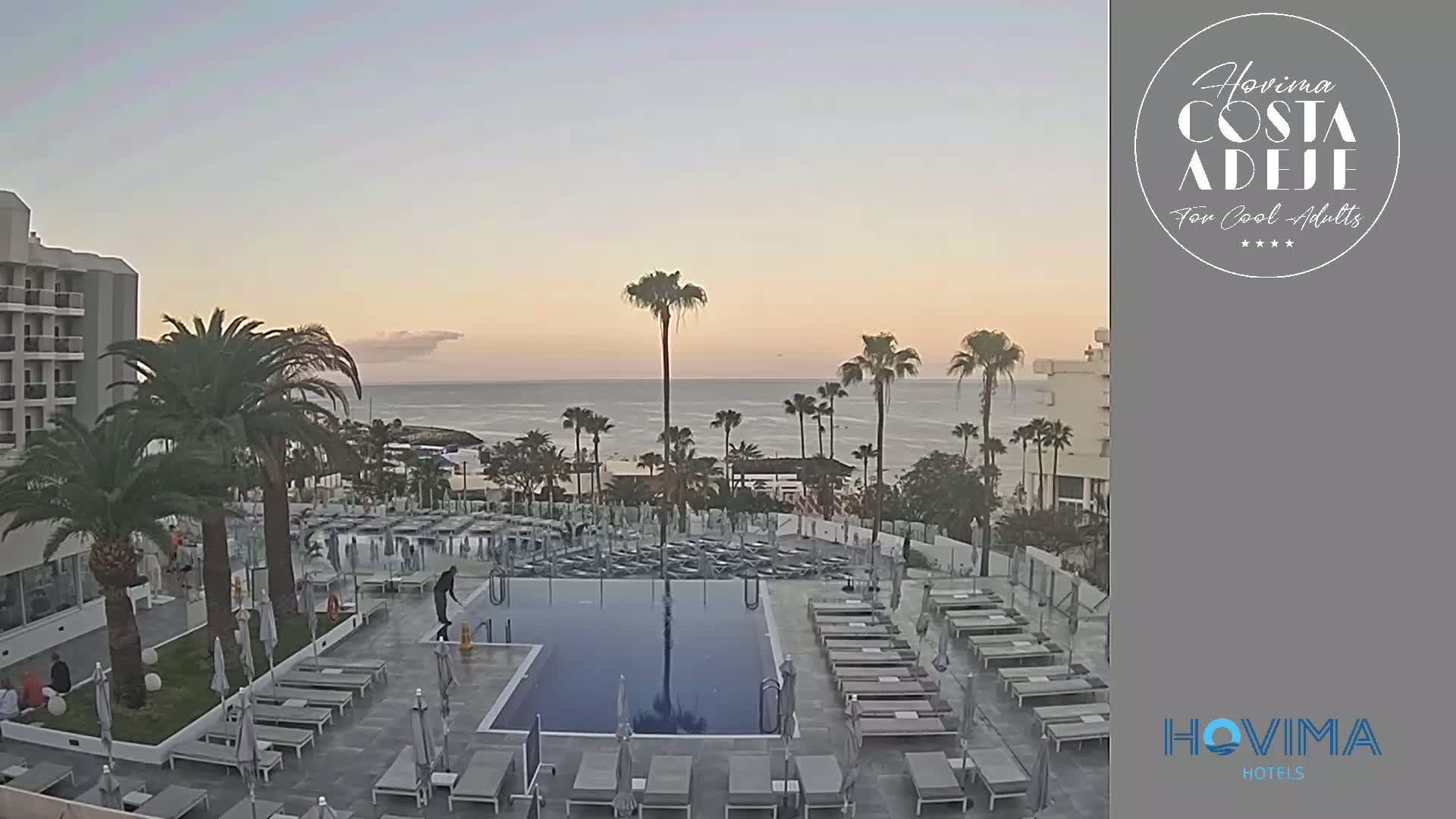 A hotel pool area with lounge chairs surrounding a rectangular pool overlooks a calm ocean under a clear, twilight sky.