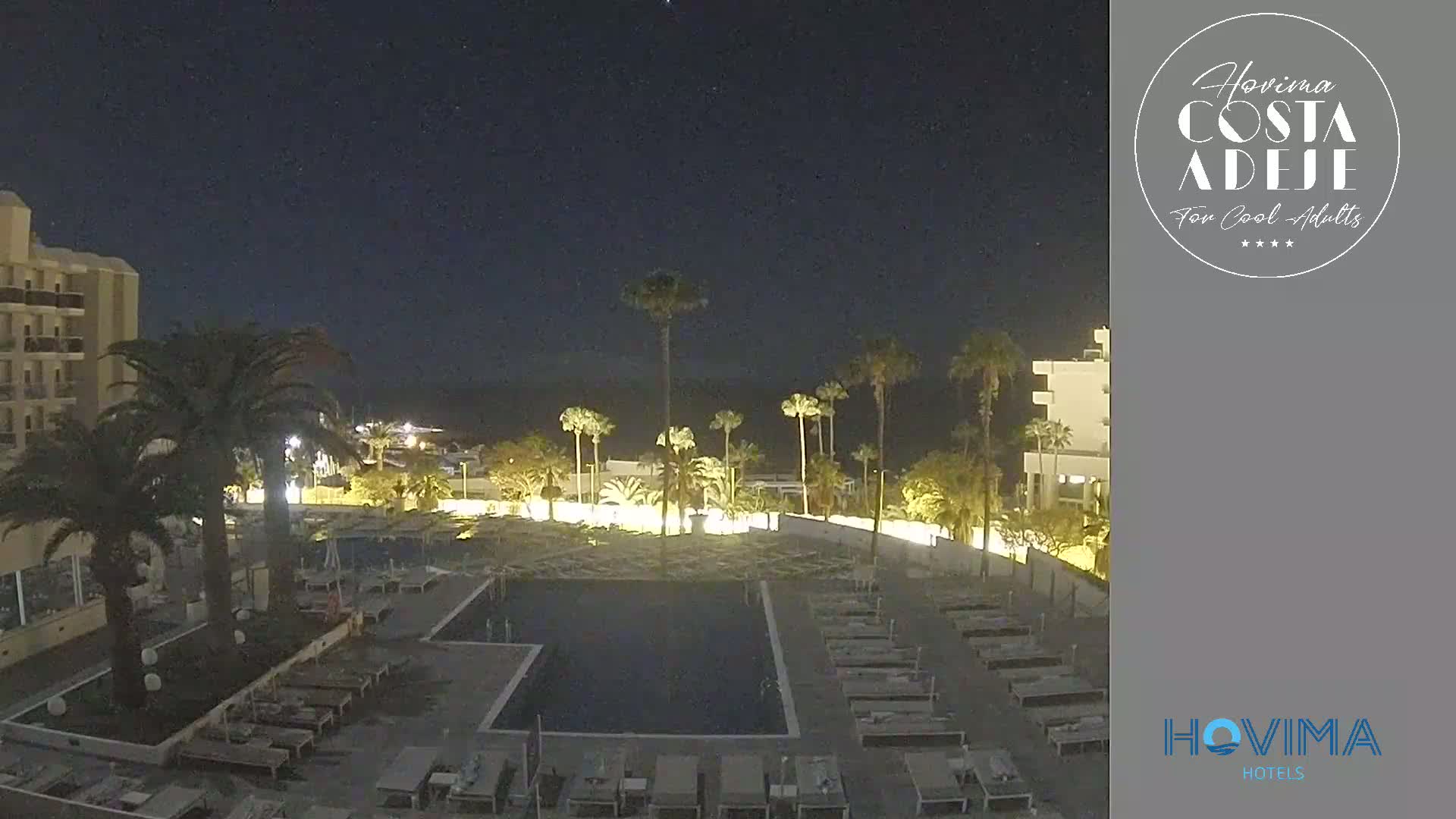 A nighttime view of a resort pool area with palm trees, surrounding buildings, and lounge chairs under a clear, dark sky.