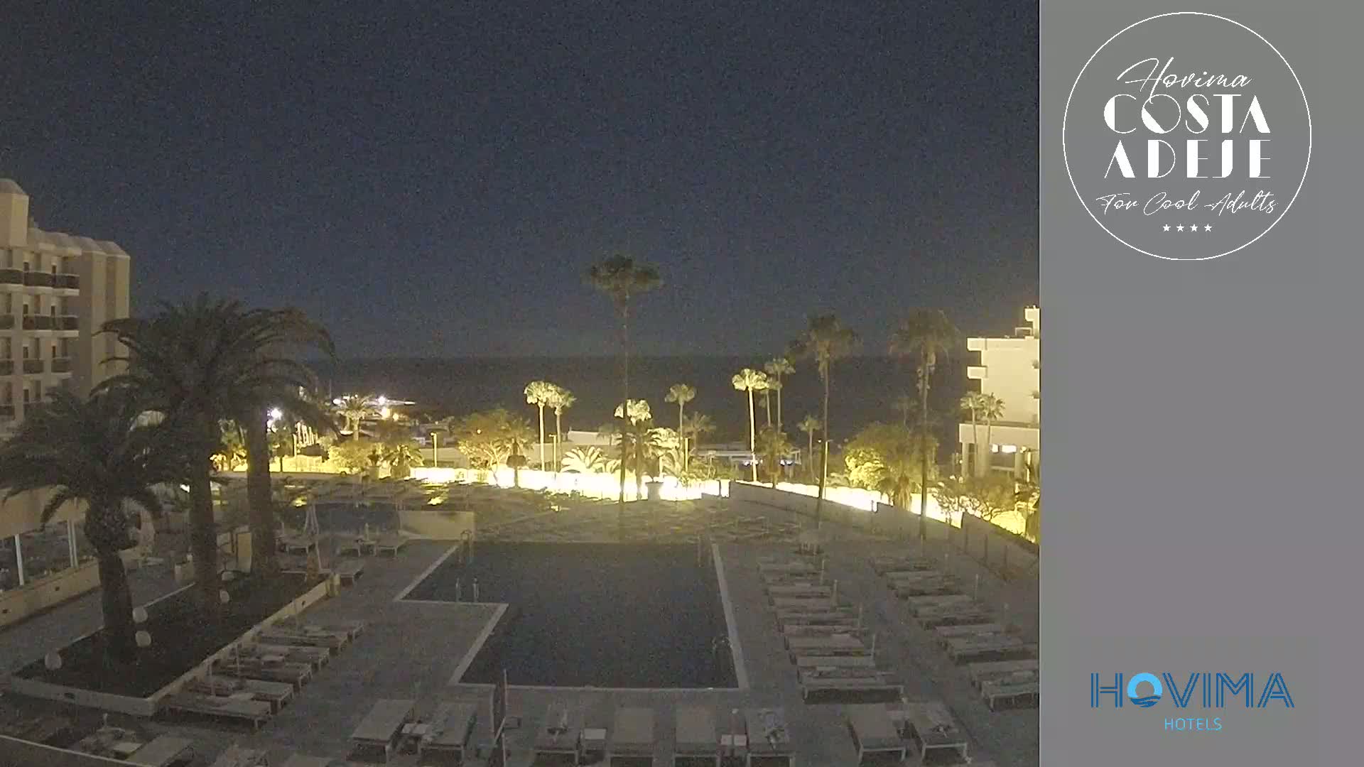 A nighttime view of a resort pool area with palm trees,  buildings, and lounge chairs under a clear, dark sky.
