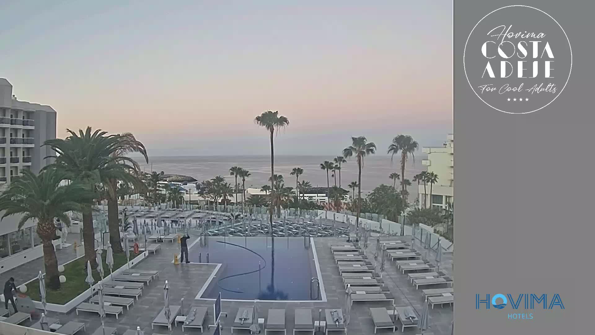 A resort pool area with numerous lounge chairs surrounding a rectangular pool, palm trees, and a calm ocean in the background under a pale, twilight sky.