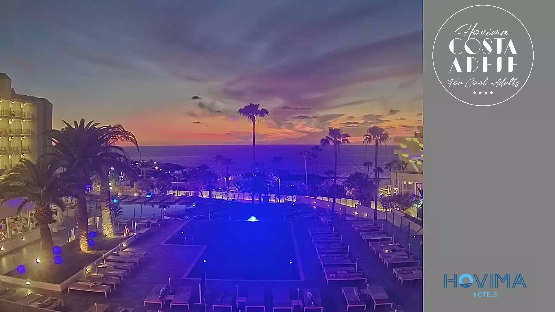An illuminated pool area with lounge chairs at a resort overlooks a calm ocean during a sunset.