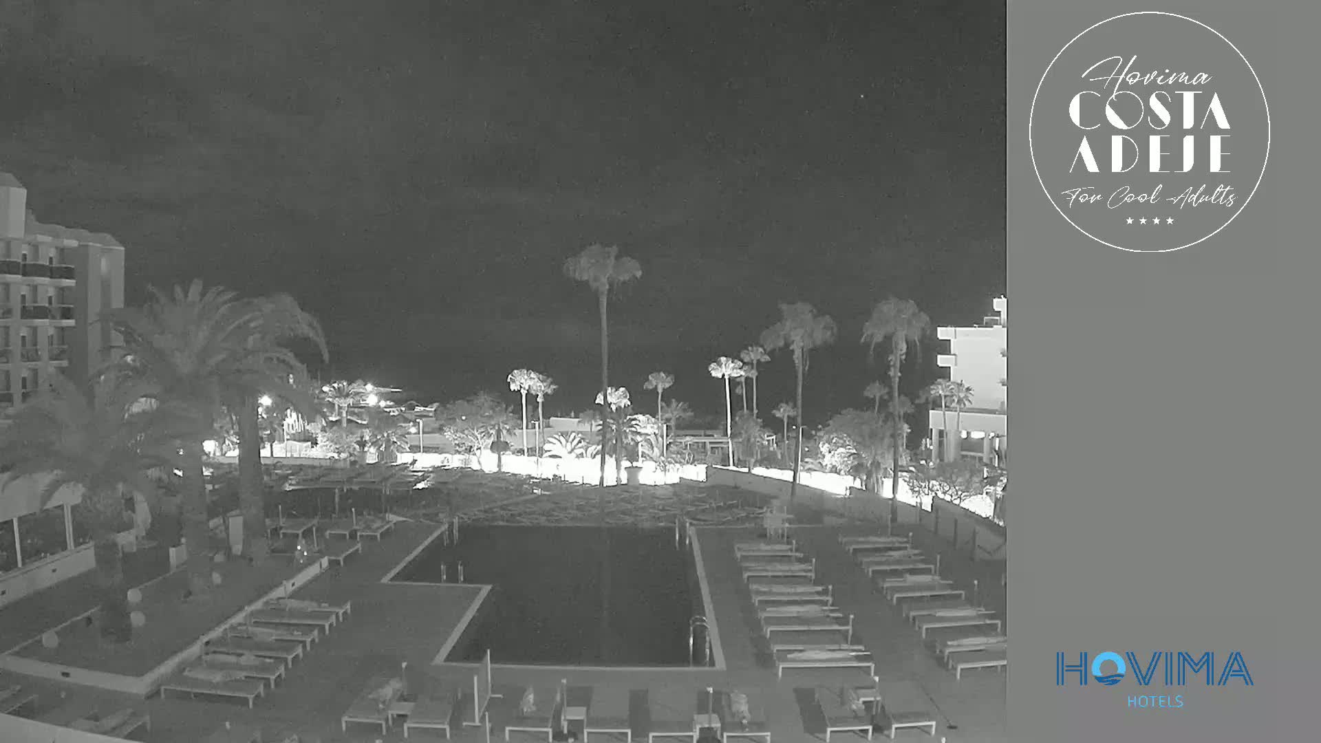 A nighttime view of a resort pool area with palm trees, lounge chairs, and a building under a clear, dark sky.