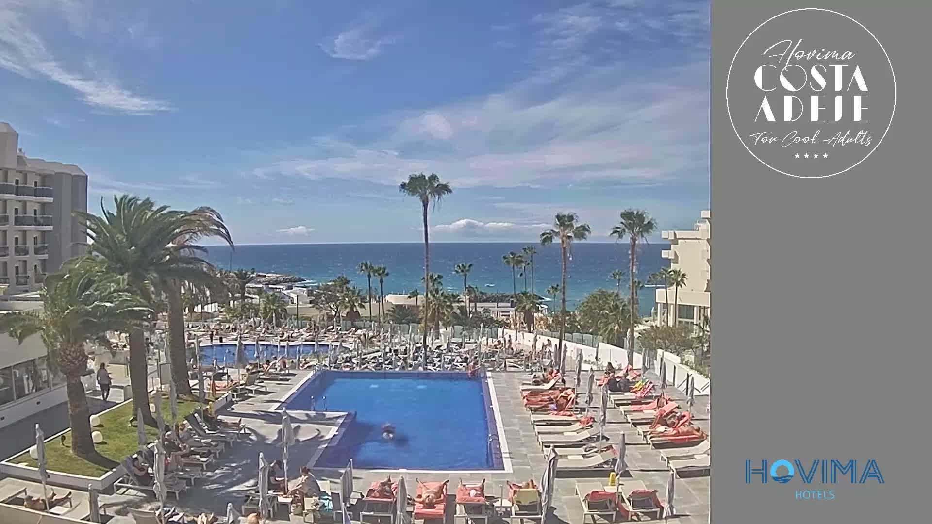 A resort pool area with many lounge chairs surrounding a large swimming pool, palm trees, and a clear ocean view under a sunny sky.