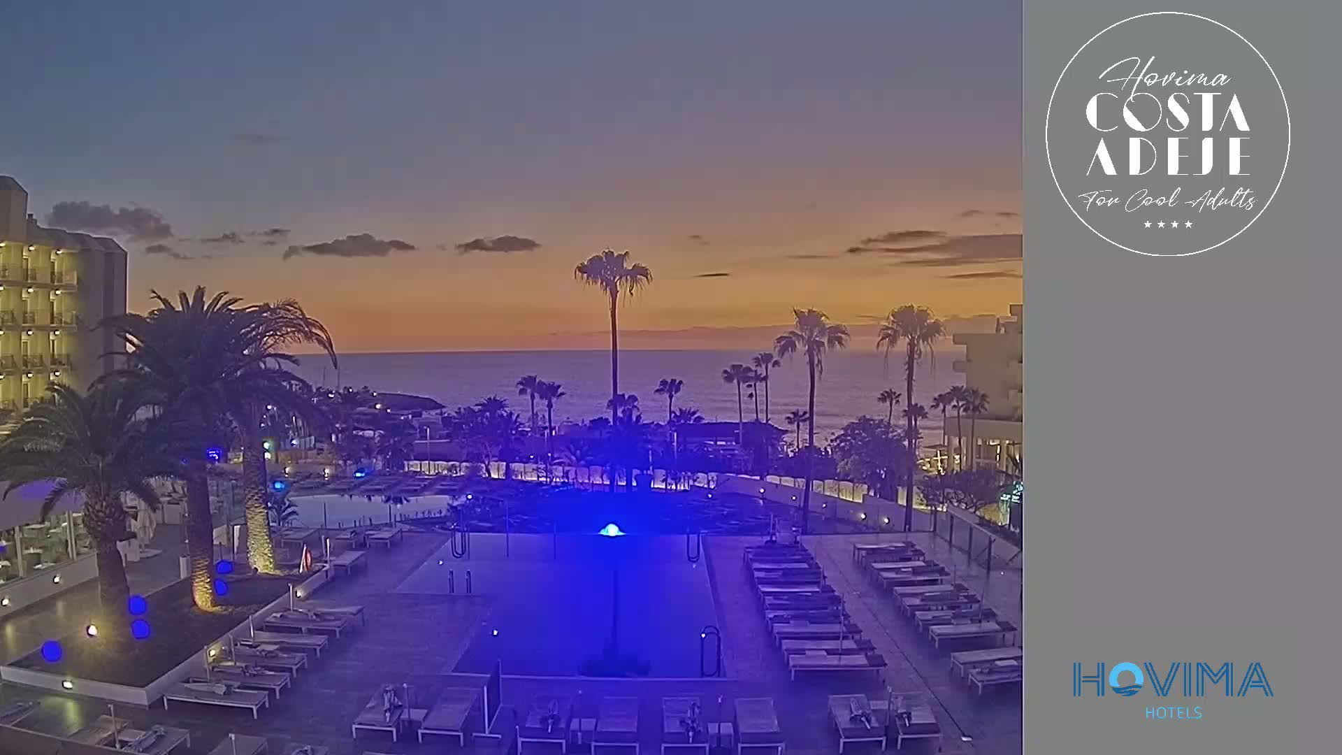 An outdoor pool area with lounge chairs is illuminated at twilight, overlooking a calm ocean under a partly cloudy sky.