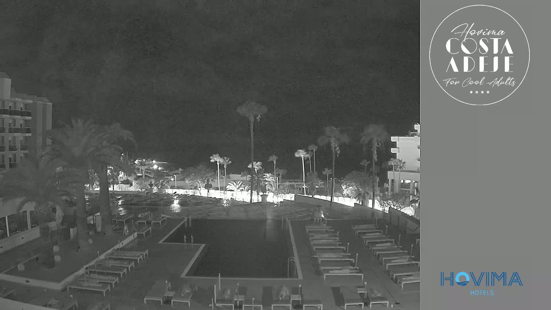 A nighttime, black-and-white view of a resort pool area with palm trees and hotel buildings under a dark, cloudy sky.