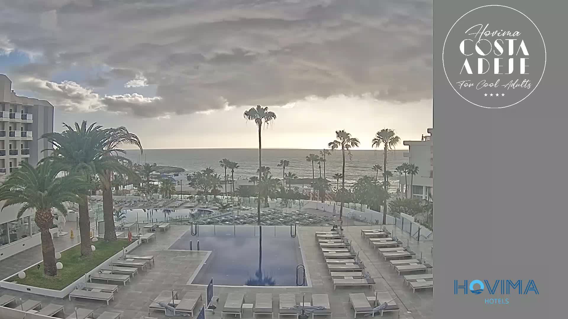An outdoor pool area with lounge chairs and palm trees is adjacent to a hotel building overlooking a calm ocean under an overcast sky.