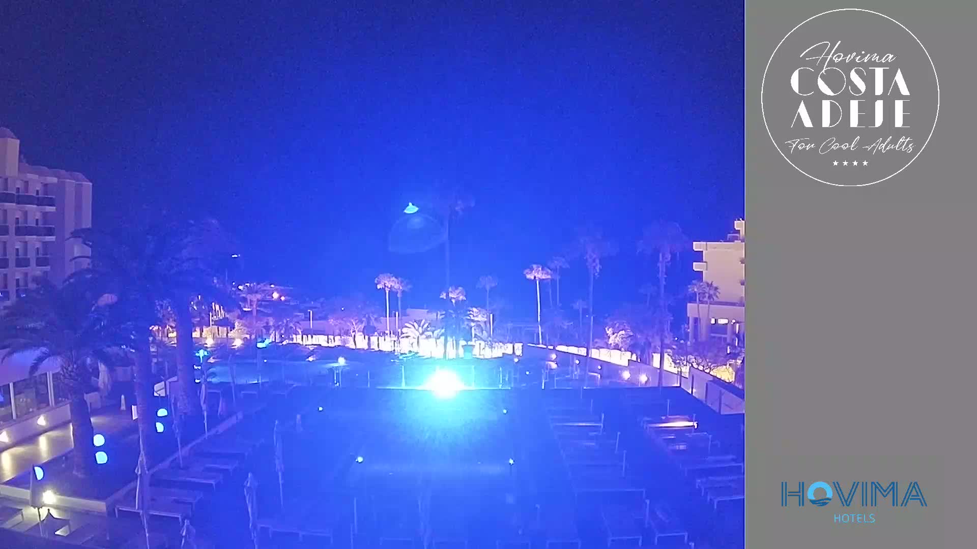 A nighttime view of a resort pool area illuminated in blue light, surrounded by palm trees and buildings under a clear, dark sky.