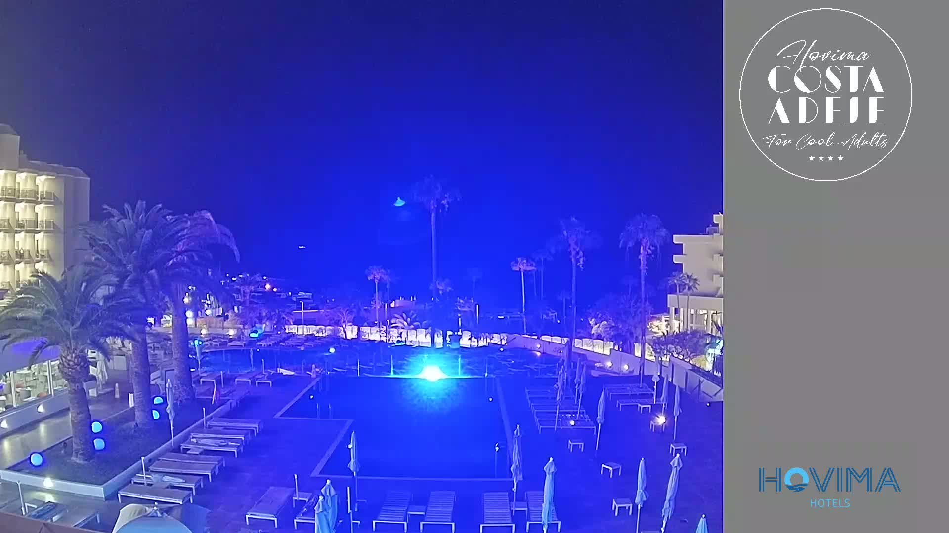 A nighttime view of a resort pool area with palm trees and a multi-story building illuminated under a dark blue sky.