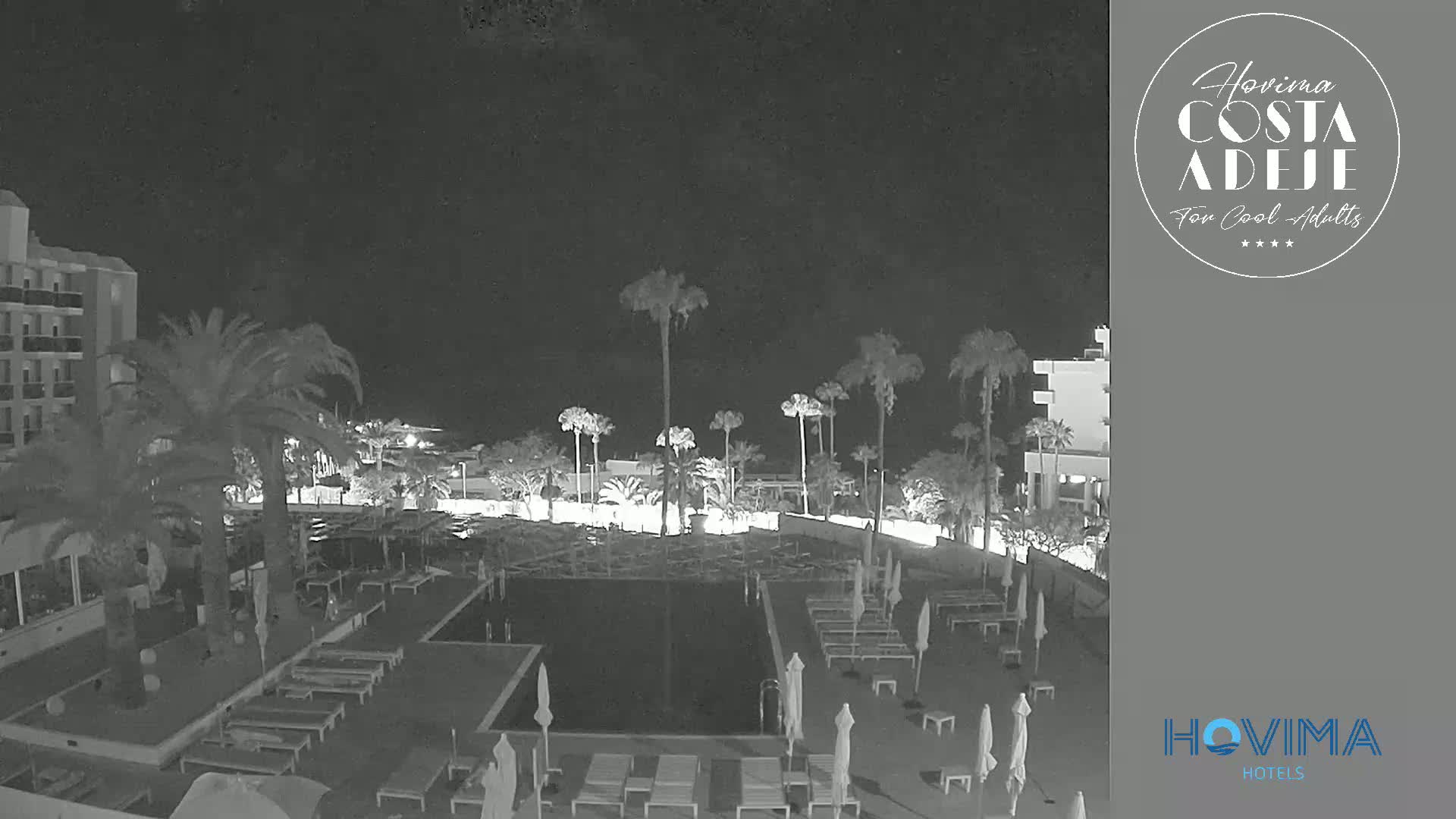 A nighttime, low-light view of a resort pool area with palm trees and surrounding buildings.