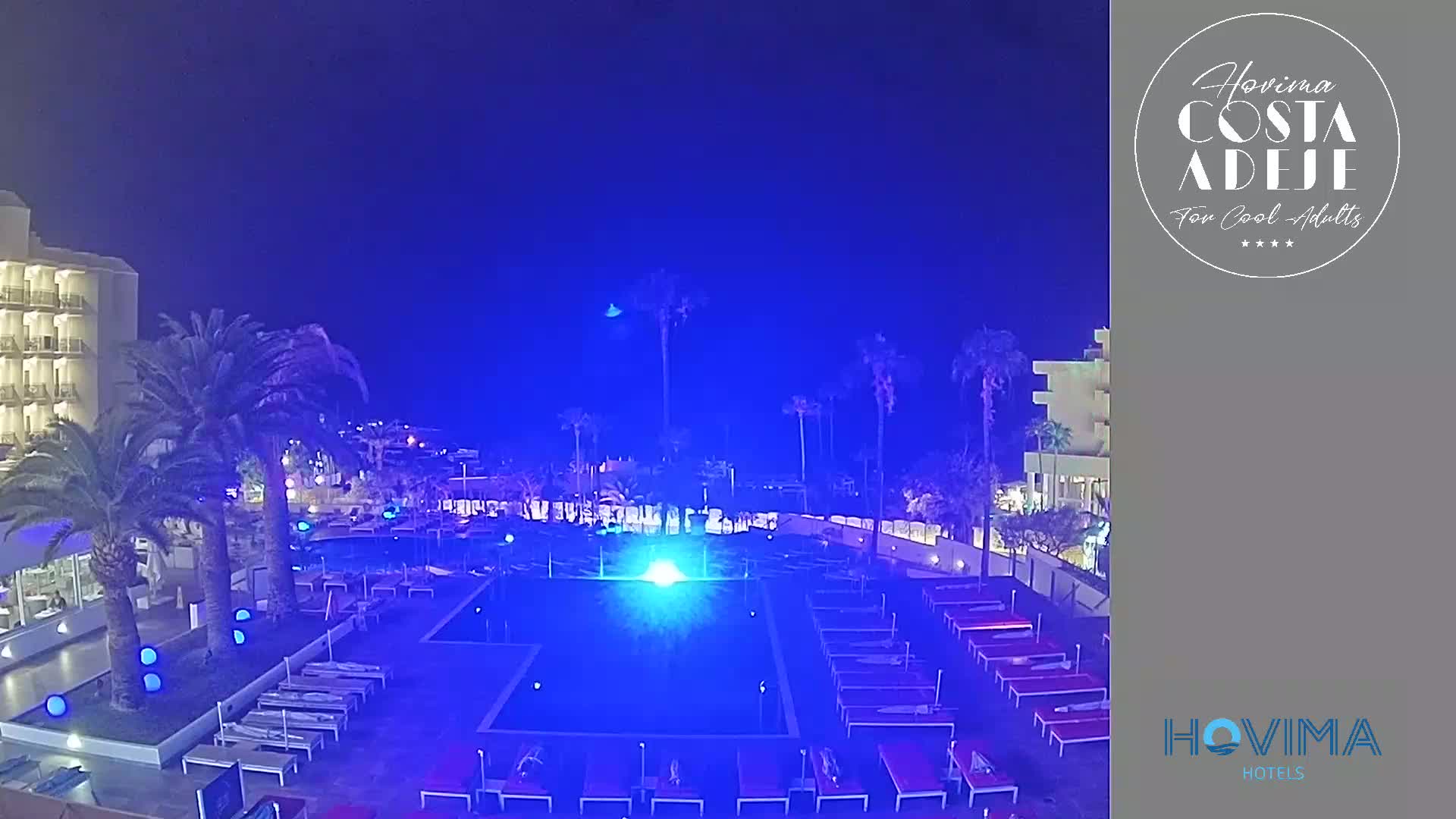 A nighttime view of a resort pool area, illuminated in blue light, surrounded by palm trees and buildings under a dark sky.