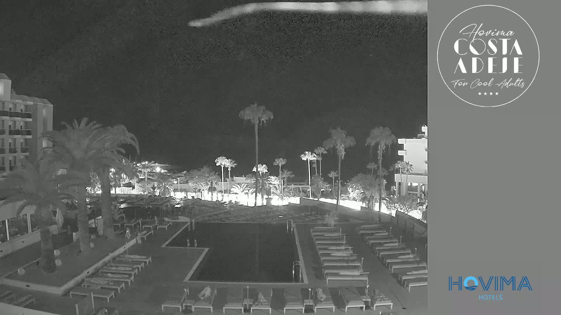 A nighttime, low-light view of a resort pool area with palm trees, lounge chairs, and a building.