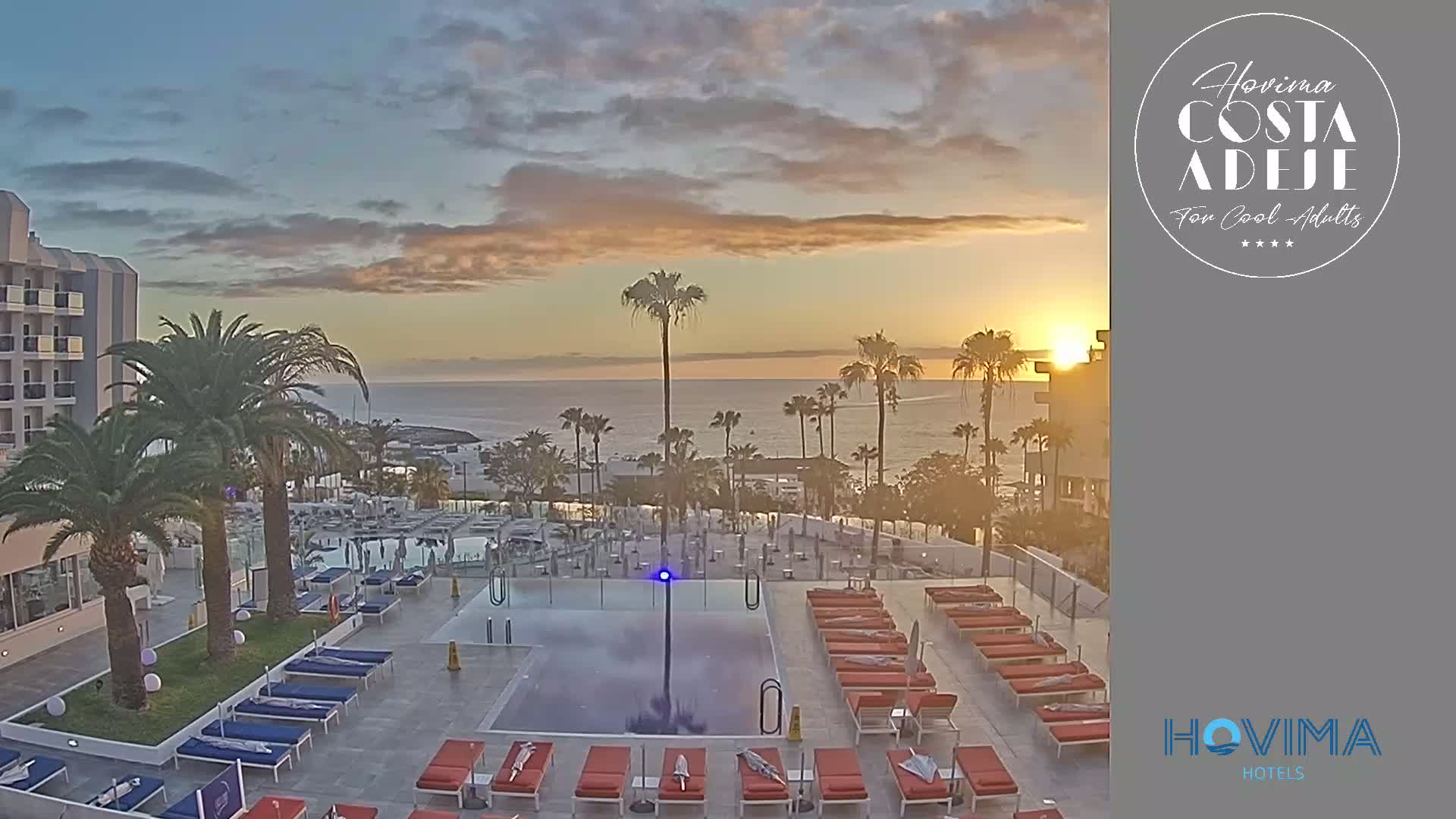 A sunrise over a calm ocean is visible from a hotel pool area with lounge chairs and palm trees.