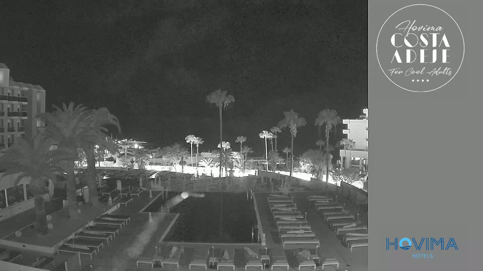 A nighttime, low-light view of a resort pool area with palm trees, lounge chairs, and a building.