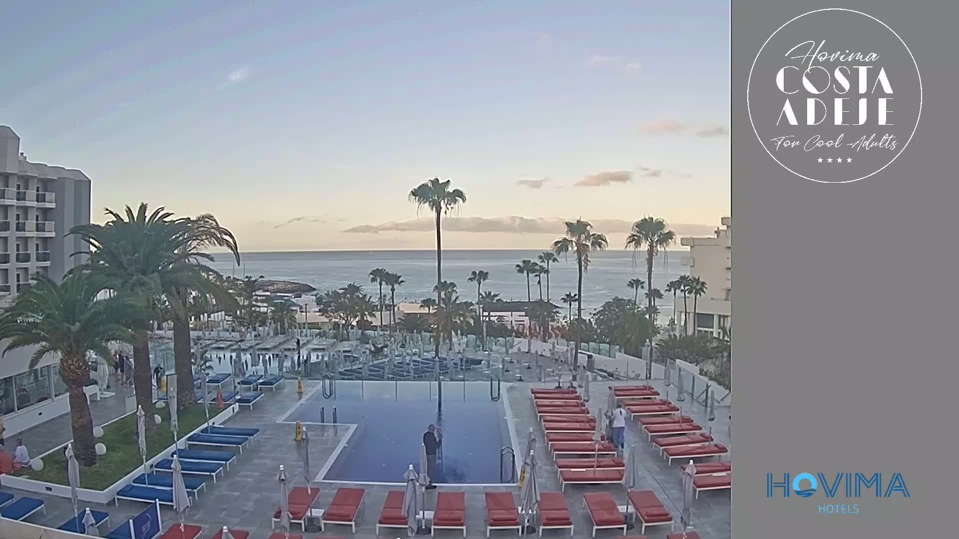 A hotel pool area with red lounge chairs surrounds a swimming pool; palm trees and a calm ocean are visible in the background under a clear, twilight sky.