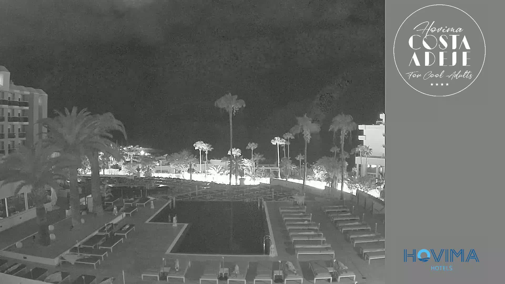 A nighttime, black-and-white view of a resort pool area with palm trees, lounge chairs, and a multi-story building.