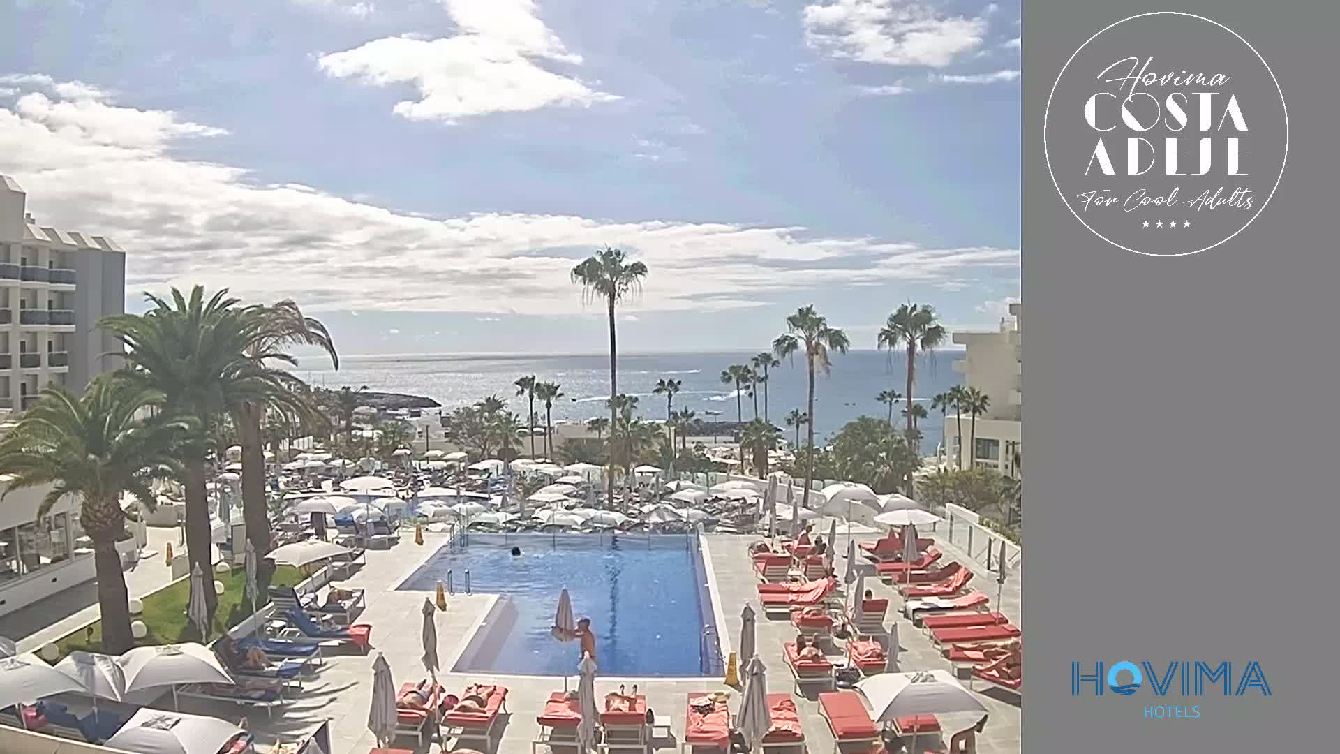 A resort pool area with red lounge chairs and umbrellas, palm trees, and a view of a calm ocean under a partly cloudy sky.