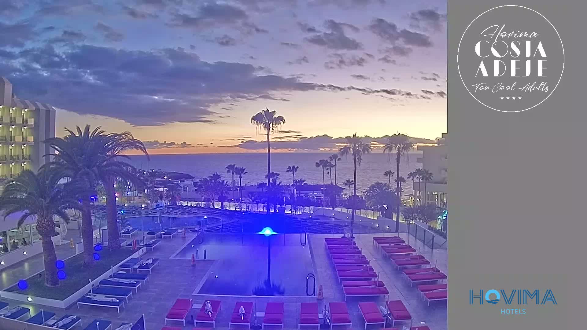 An outdoor pool area with lounge chairs, palm trees, and a building overlooks a calm ocean at sunset under a partly cloudy sky.