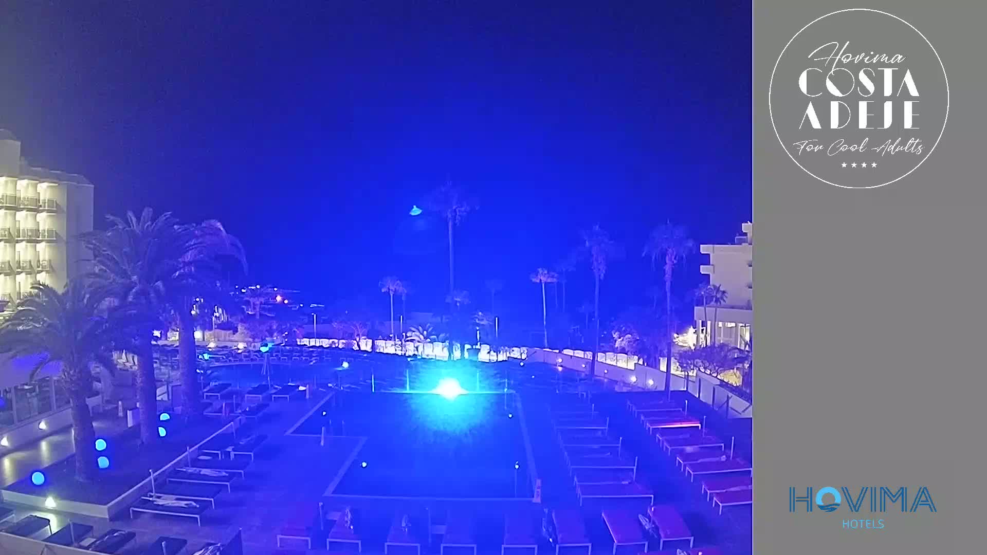 An illuminated swimming pool and surrounding lounge chairs at night, next to a hotel building and palm trees under a dark blue sky.