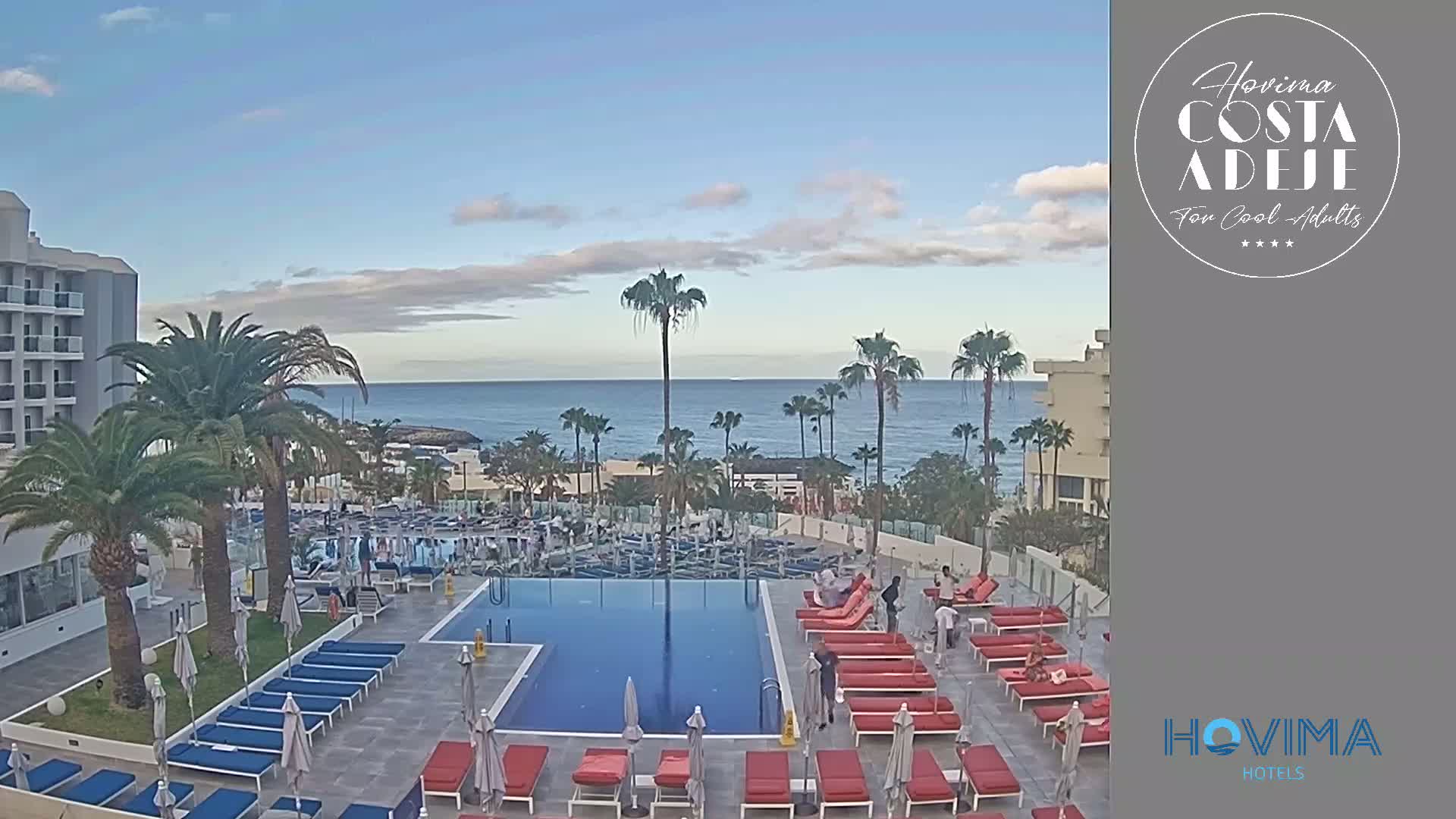 A hotel pool area with red and blue lounge chairs surrounds a rectangular pool, overlooking a calm ocean under a partly cloudy sky.