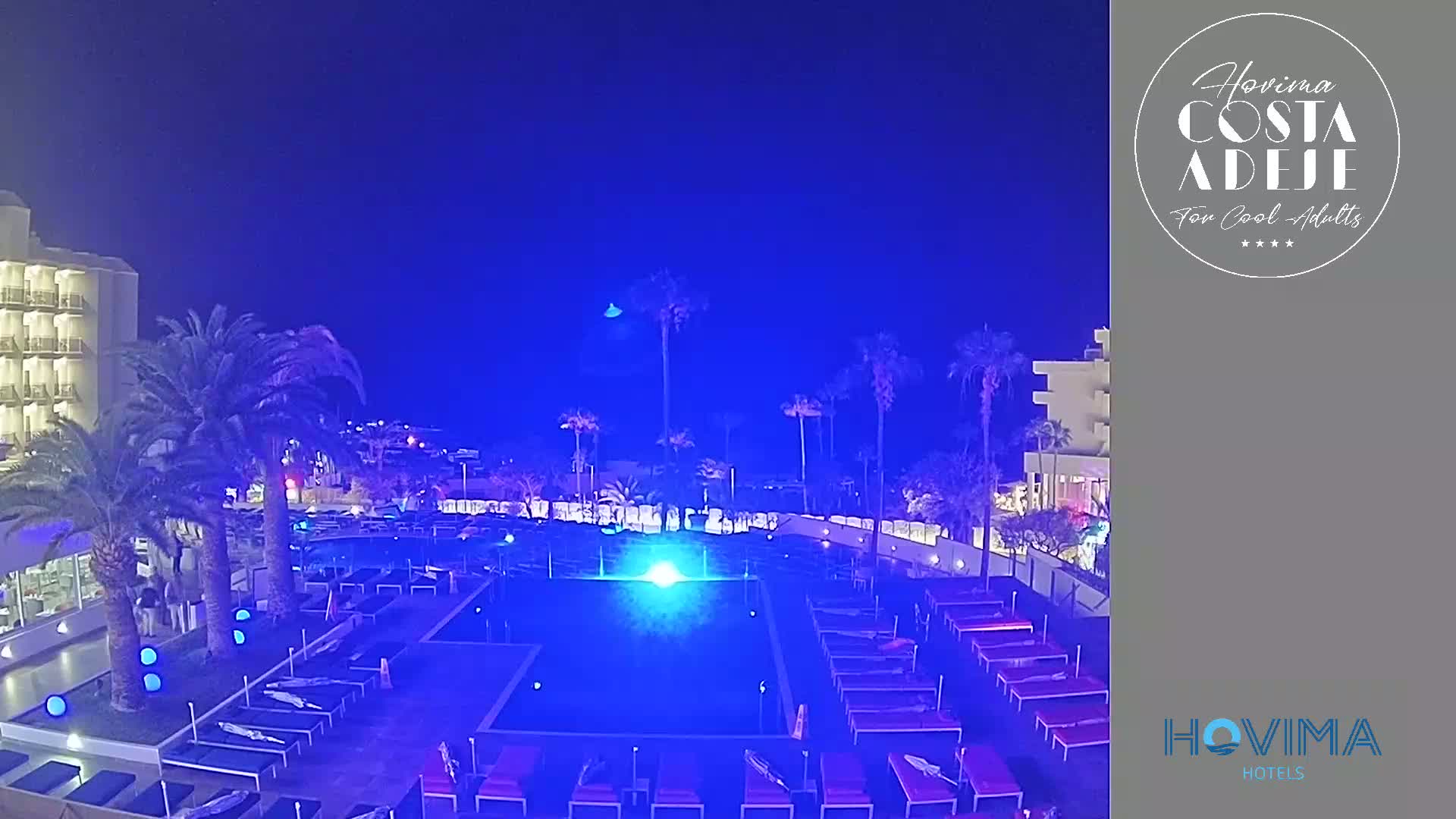 A nighttime view of a resort pool area illuminated in blue, surrounded by palm trees and a hotel building under a dark, clear sky.